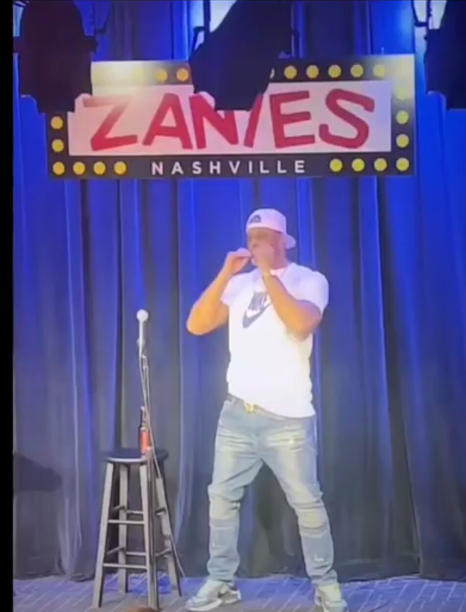 Person standing on stage with a microphone, wearing a white cap and shirt, in front of a Zanies Nashville sign.