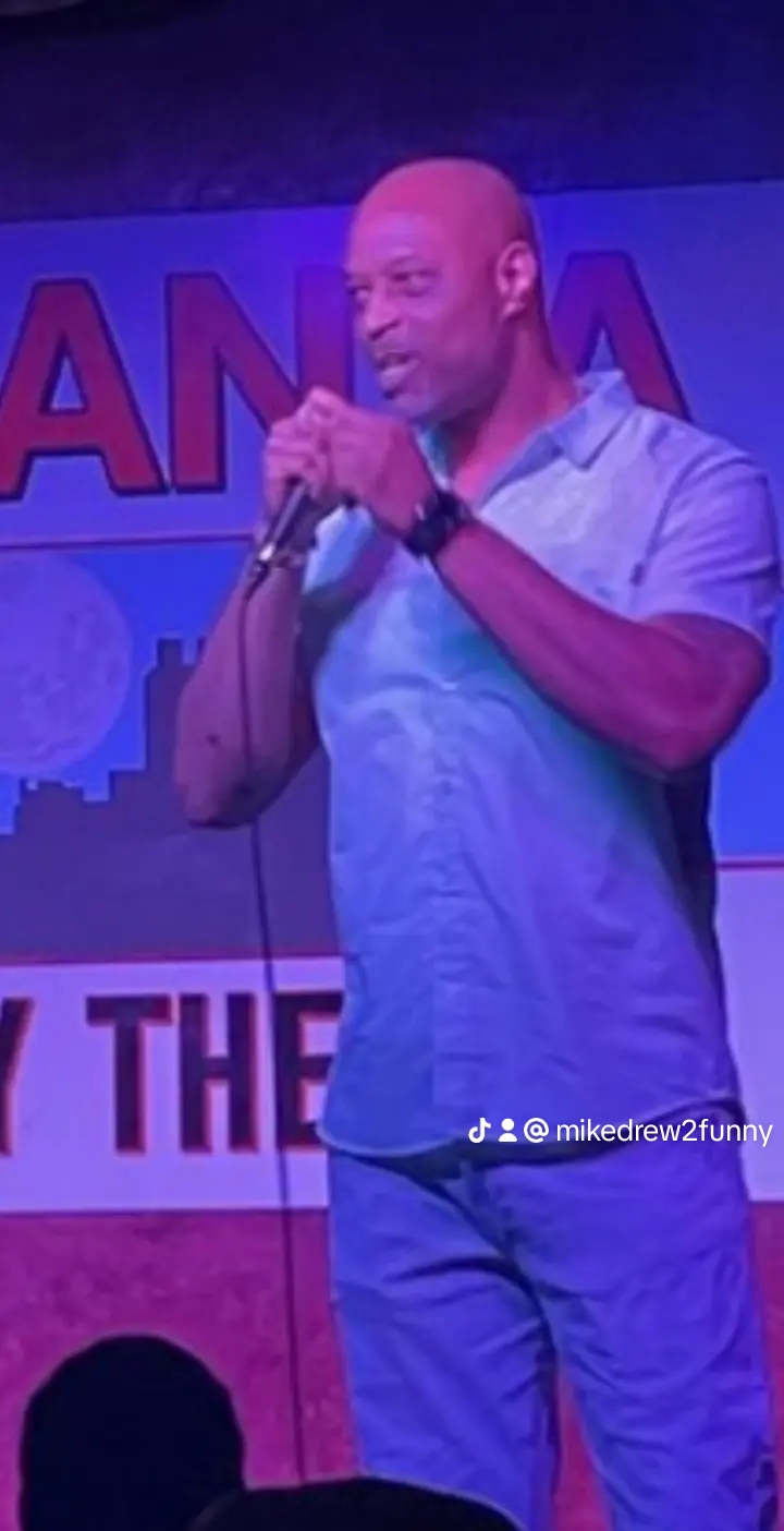 Man standing on stage holding a microphone, wearing a light blue shirt and dark pants, with a colorful background.