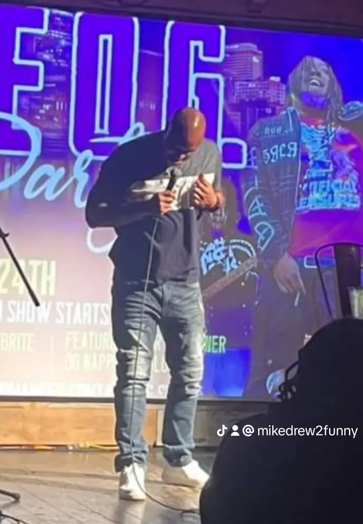Person standing on stage reading from a paper, with a large colorful background and a person in a hoodie on screen behind.