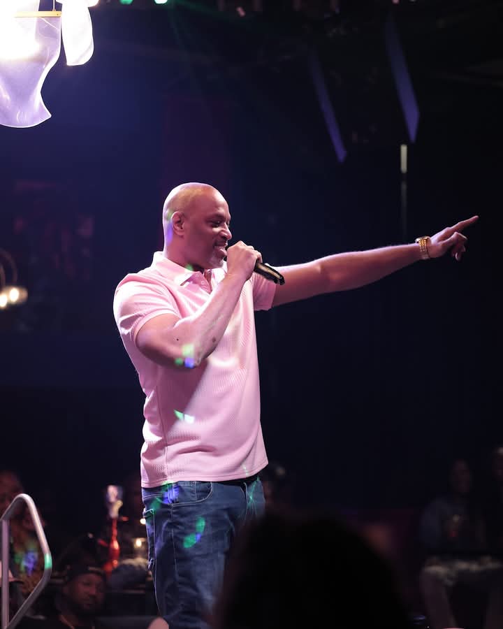 Man with a microphone pointing to the right, wearing a pink shirt and jeans, on stage with dark background.