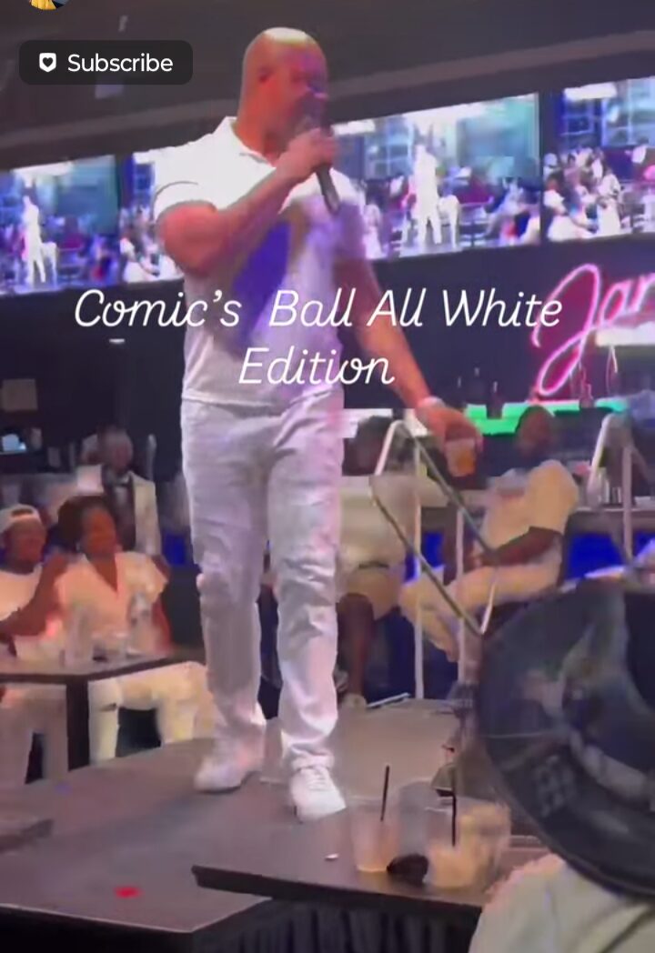 Person standing on stage speaking into microphone in a crowded indoor venue with screens and audience.