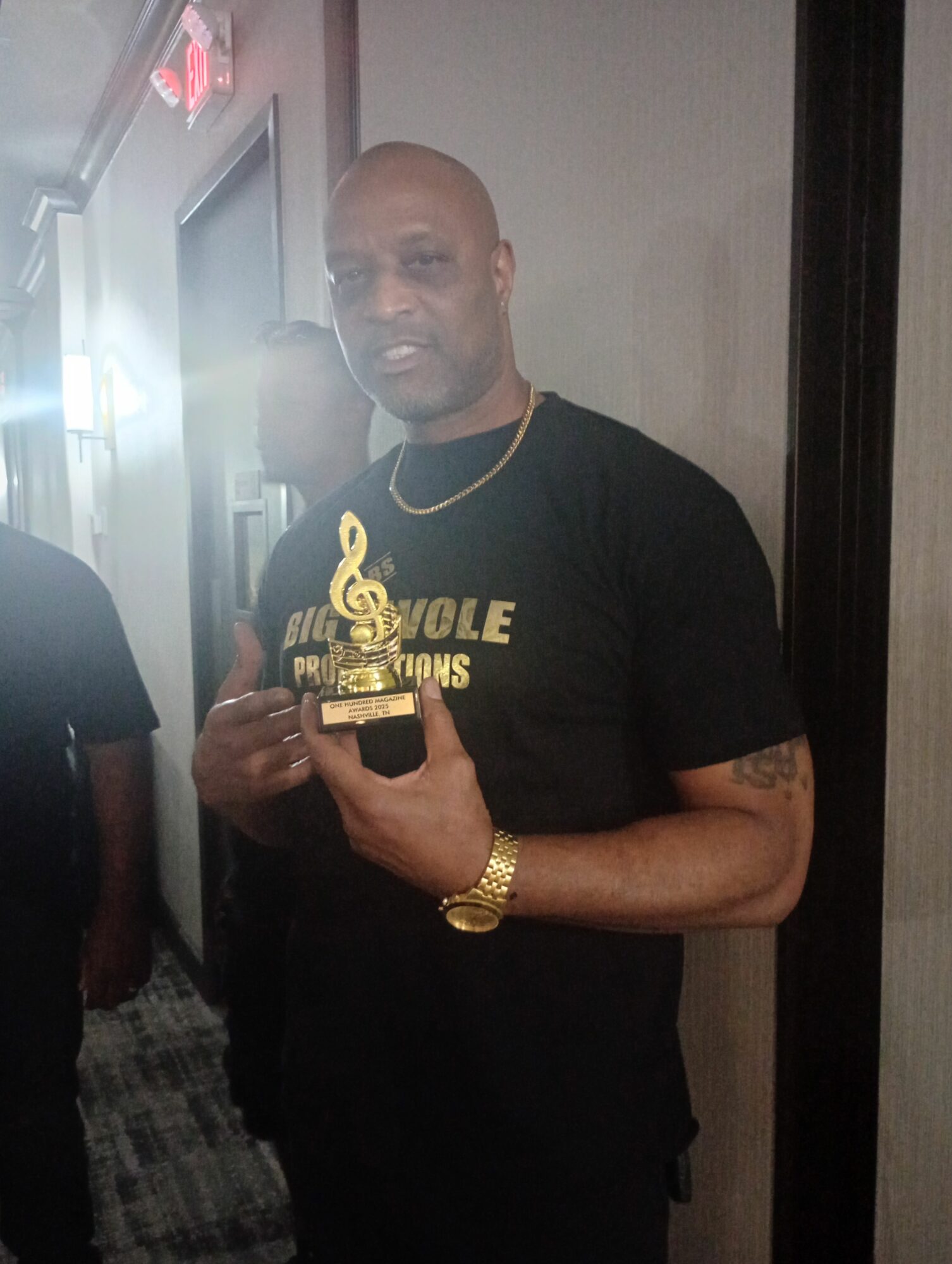 Man taking a mirror selfie, wearing a black T-shirt, gold watch, and necklace, holding a phone, standing indoors.