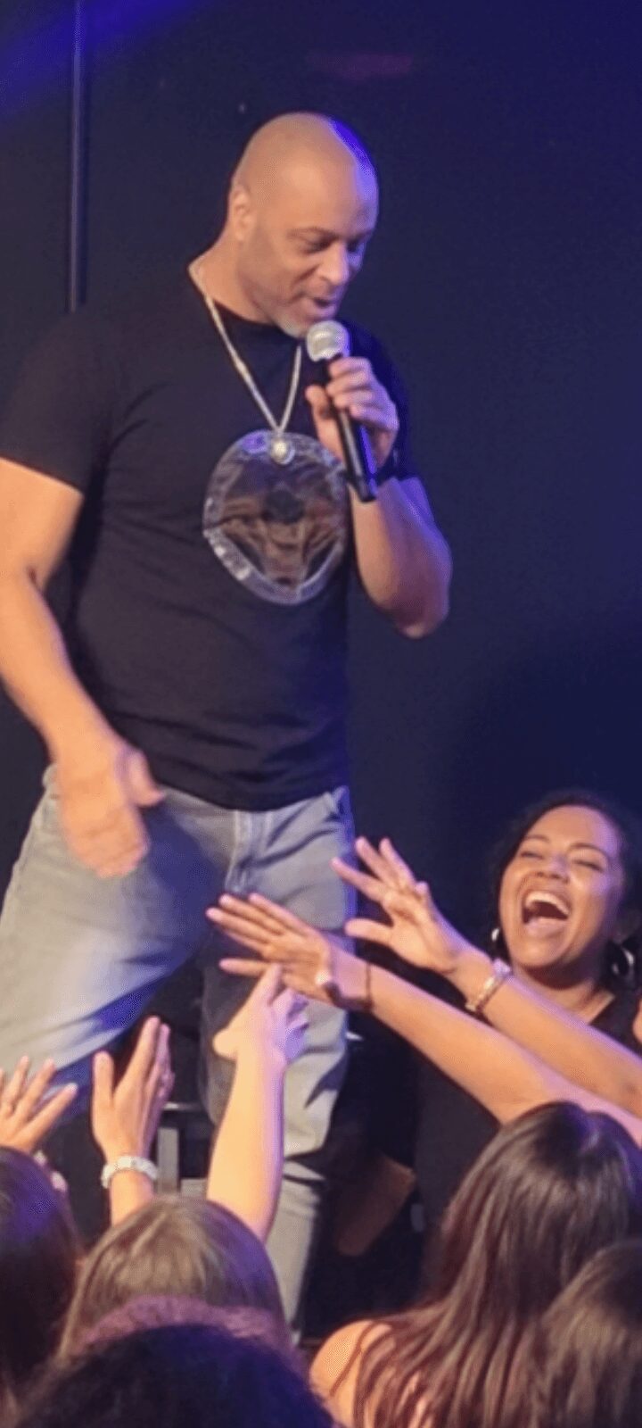 Man holding microphone on stage, smiling, audience reaching out, woman laughing nearby, dark background