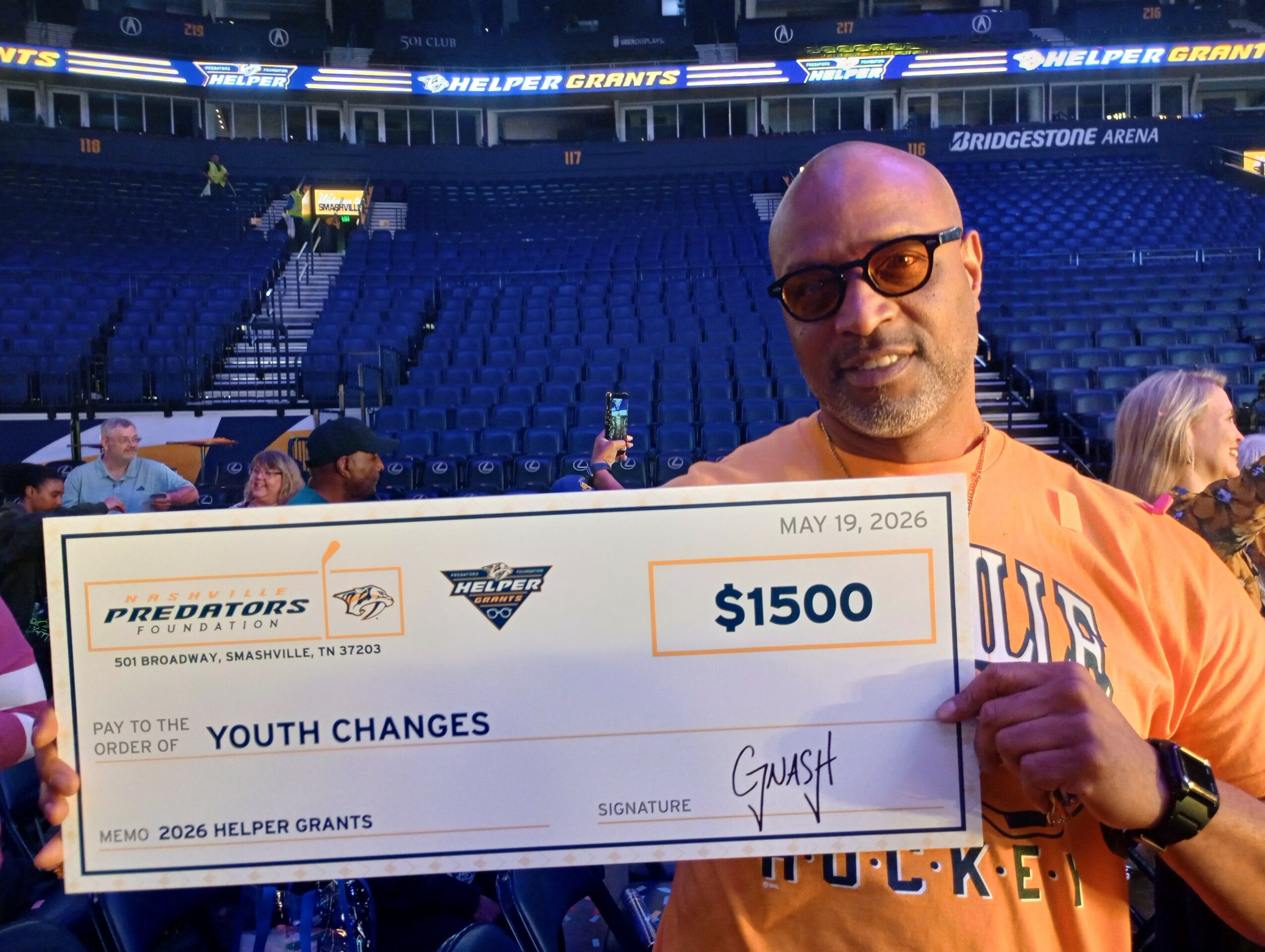 Man in orange shirt holding a large check for $1500 at a stadium, with empty blue seats in the background.