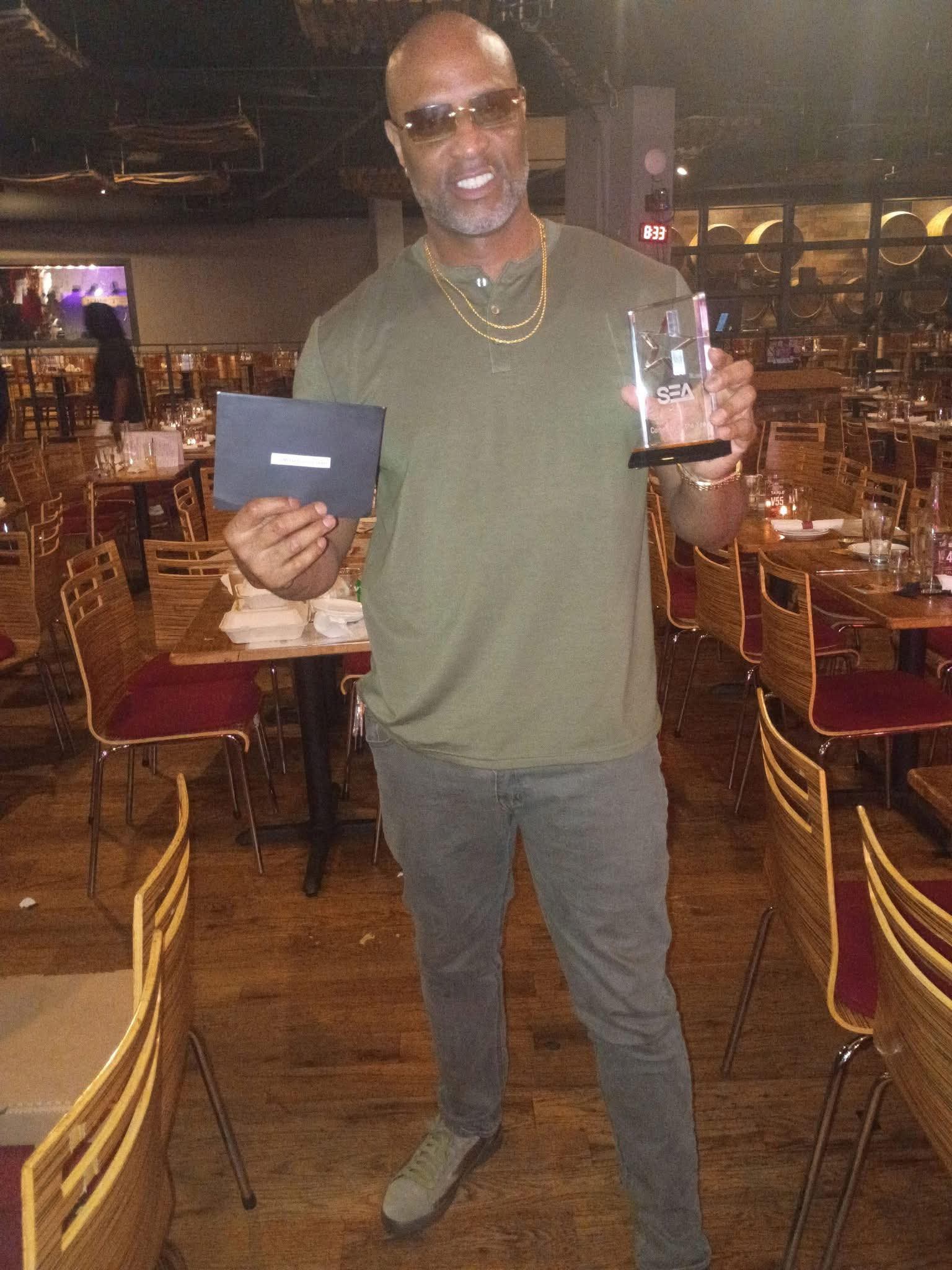 Man holding a glass award and a black box in a restaurant with wooden chairs and tables.