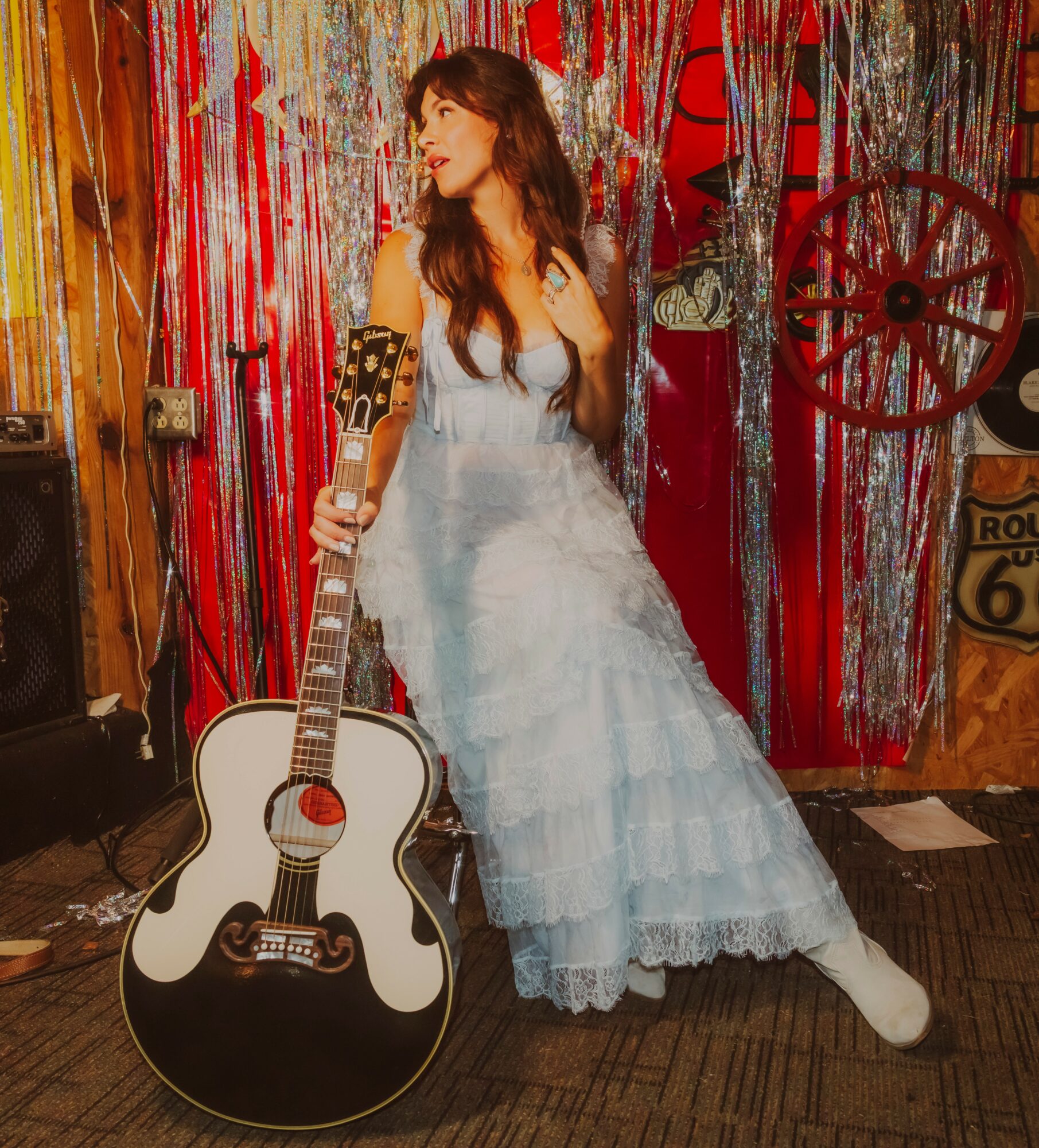 Young woman in a light blue dress holding a guitar, standing in a decorated room with red and silver streamers.