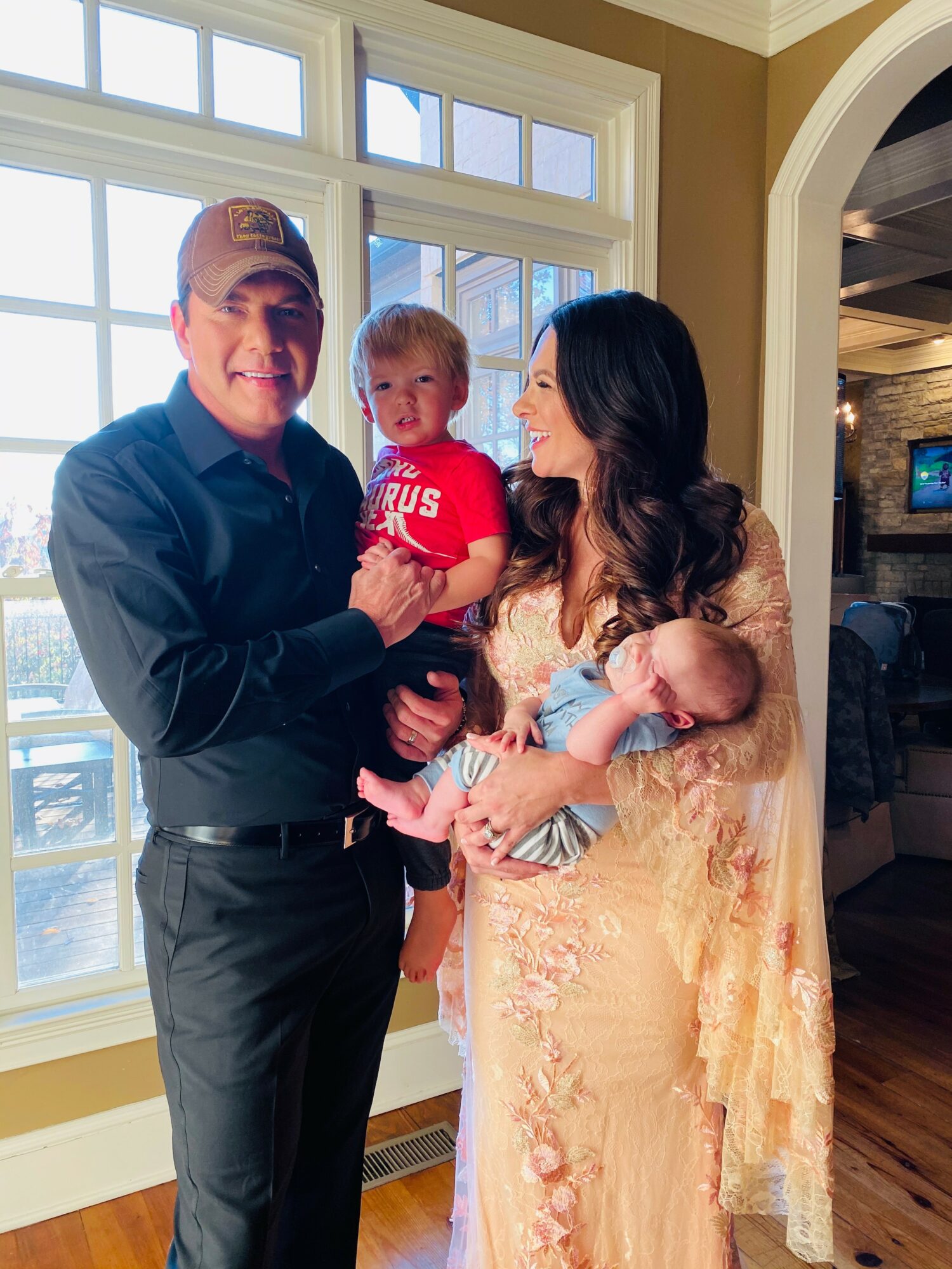 Family of three with a man, woman, and child standing indoors near a large window, smiling and holding the child.