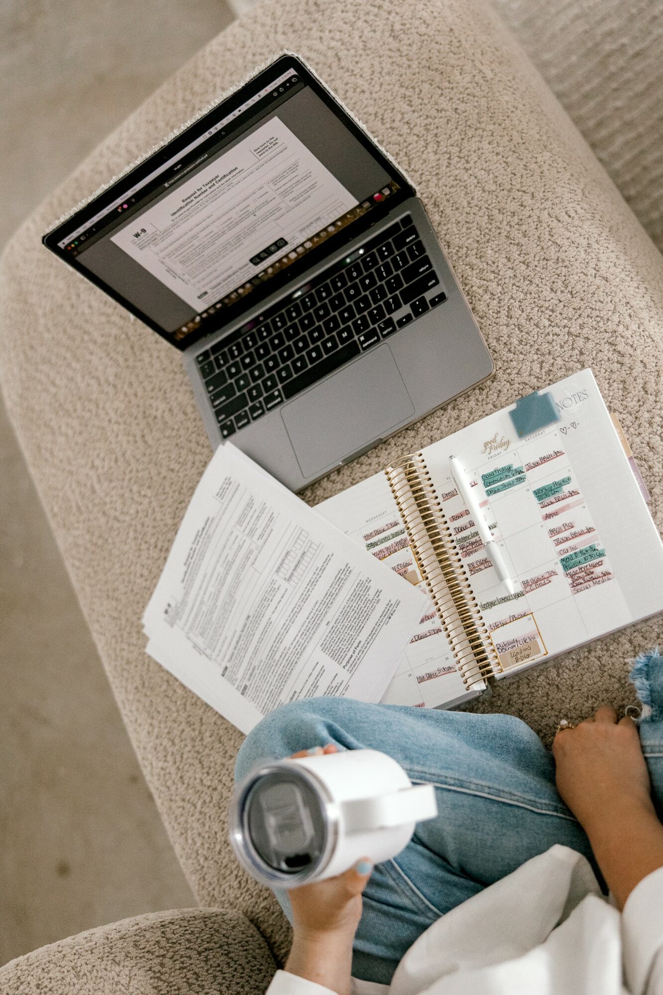 Laptop, papers, and a person holding a white device on a beige couch.