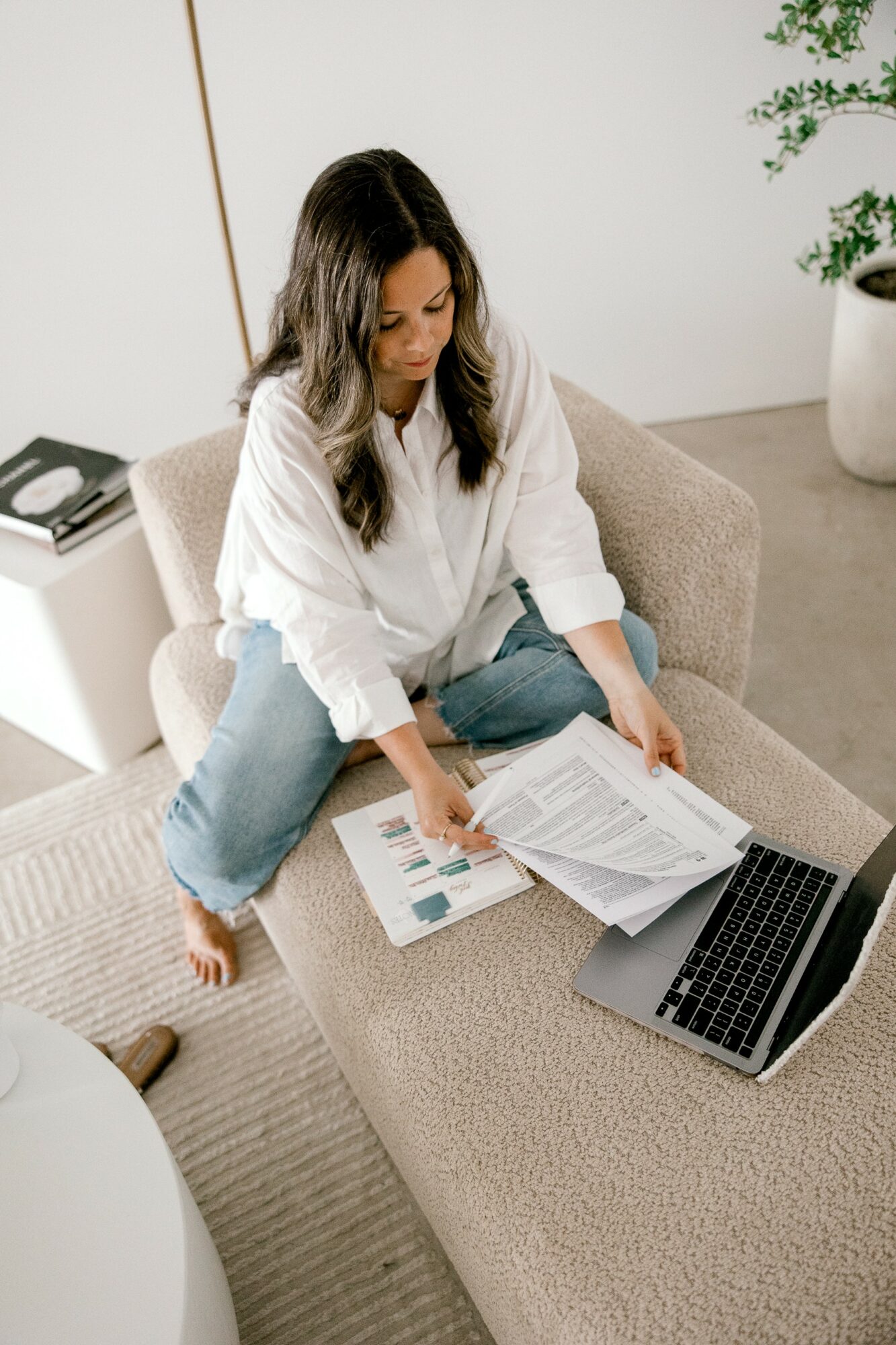 Woman sitting on a sofa, reviewing documents with a laptop open nearby, in a bright room with a plant.
