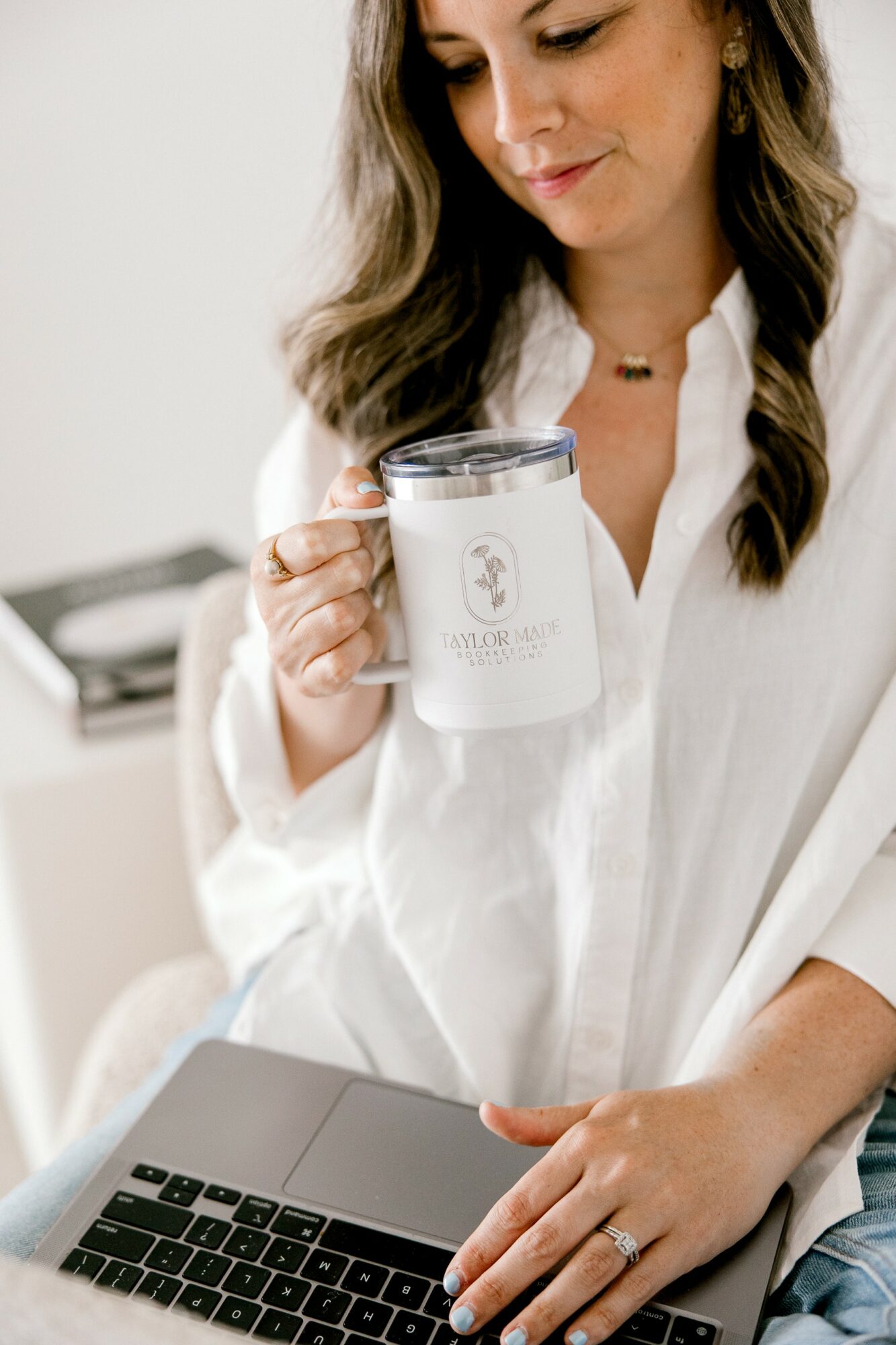 Woman holding a white mug with a logo, working on a laptop, wearing a white shirt, in a bright room.