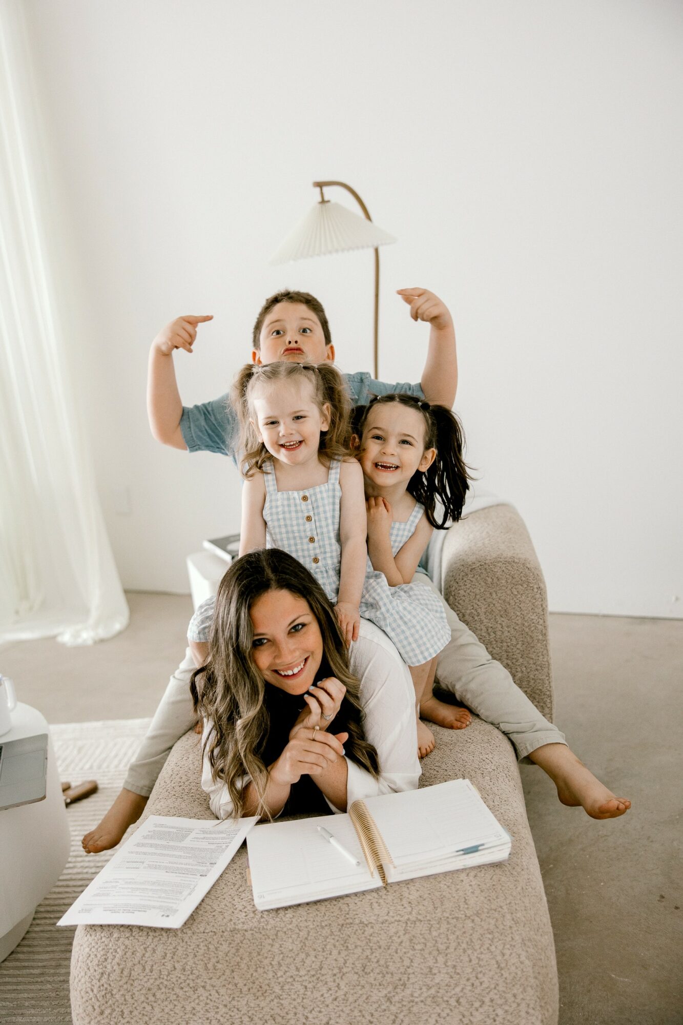 Four children sitting on a beige sofa, smiling and reading books, with a white wall and lamp behind them.