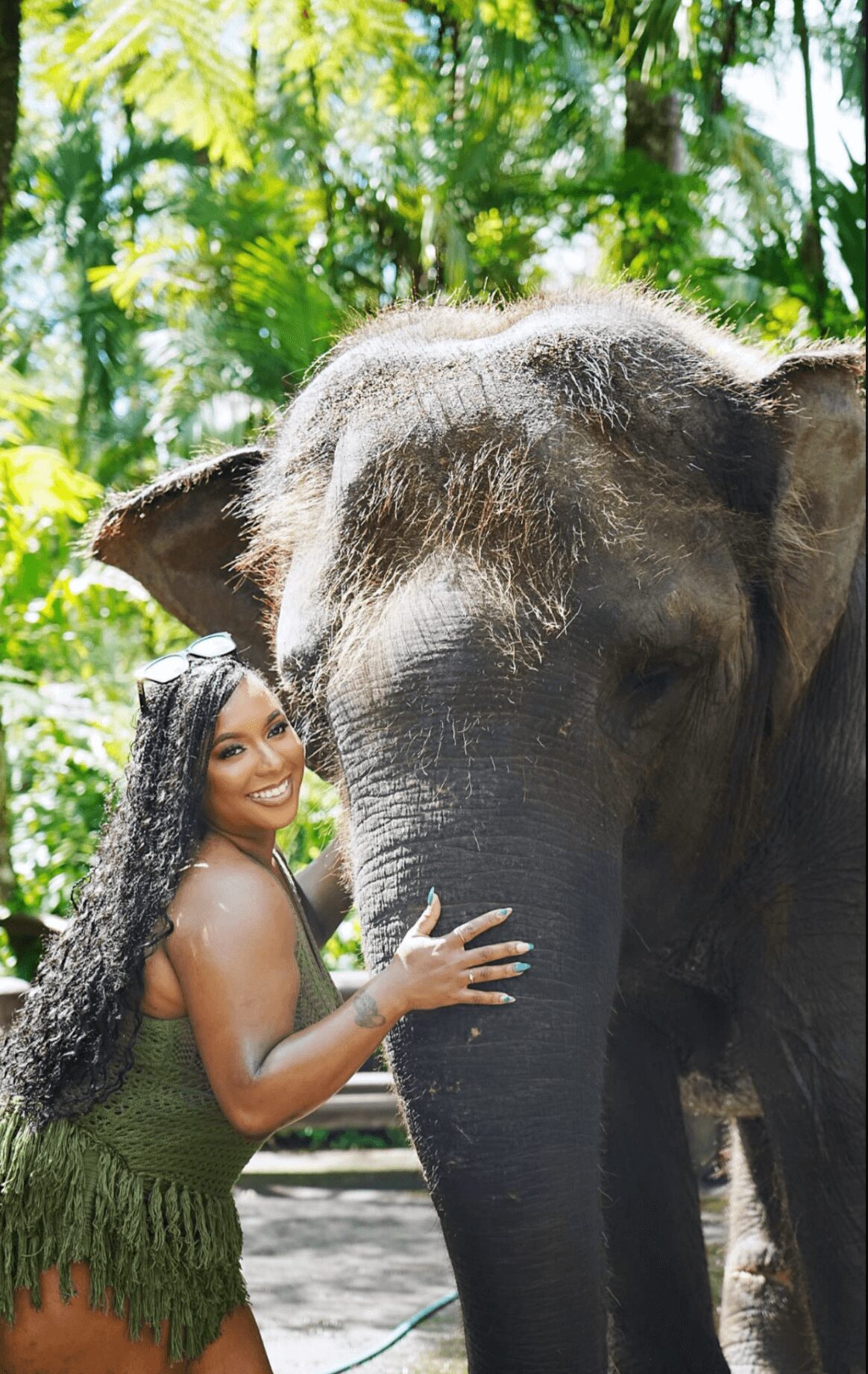 Young woman with long curly hair touching an elephant's face outdoors in a lush green environment.
