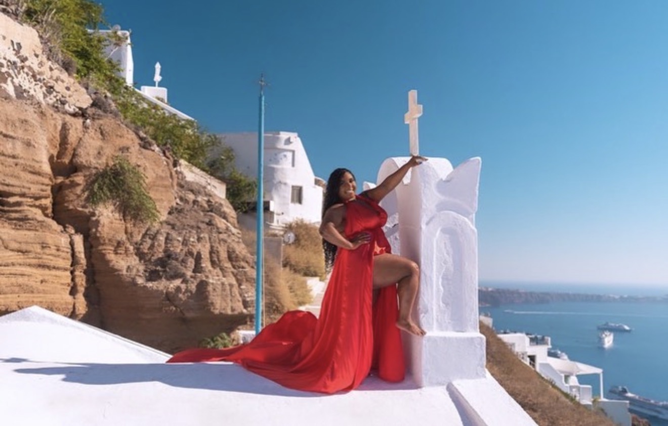 Woman in red dress sitting on white structure near a cross, with cliffs and sea in background.