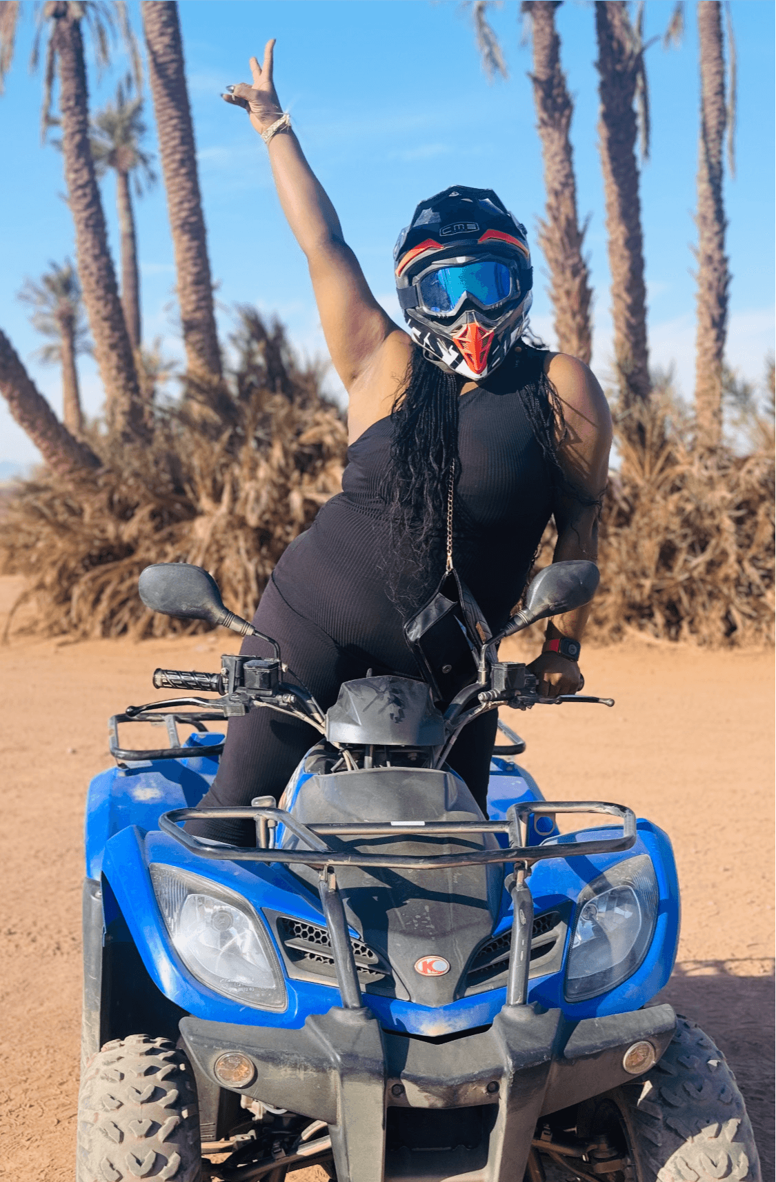 Person wearing a helmet and black outfit, sitting on an ATV with palm trees in the background, raising one arm.