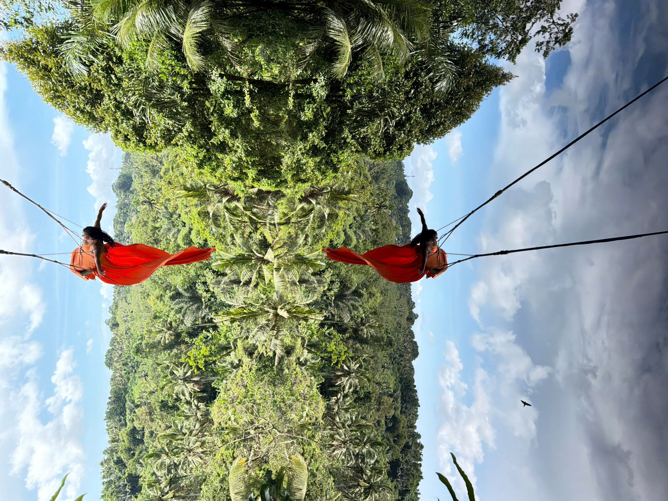 Two people walk on a tightrope over a river with lush green trees and a partly cloudy sky.