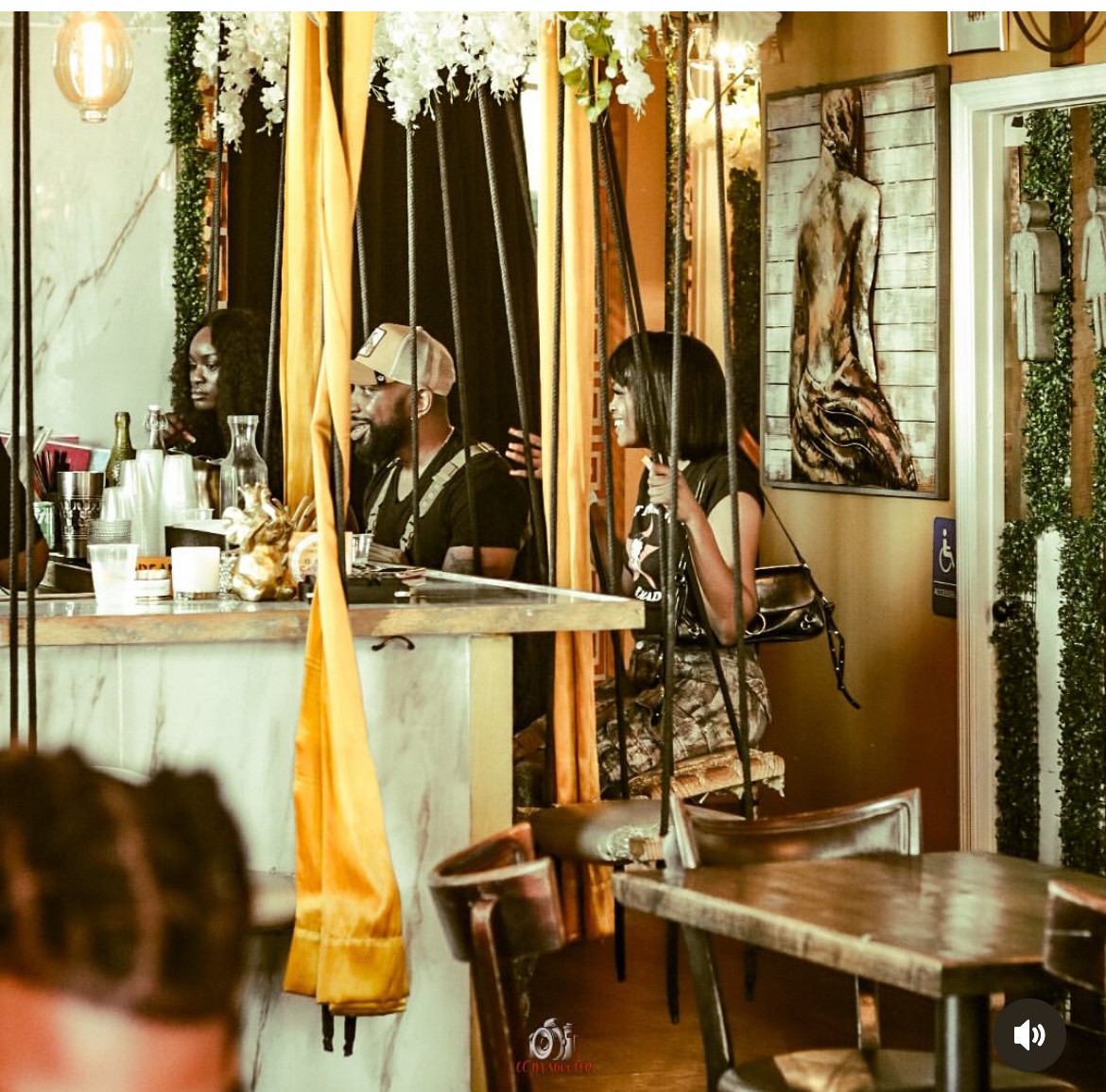 People sitting at a bar counter in a decorated indoor space with hanging flowers and artwork on the wall.