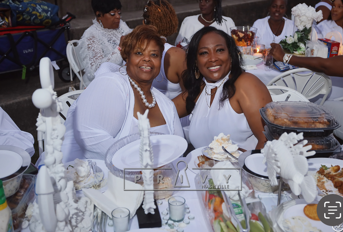 Two women smiling at a table during a celebration, surrounded by food and decorations, with other guests in the background.