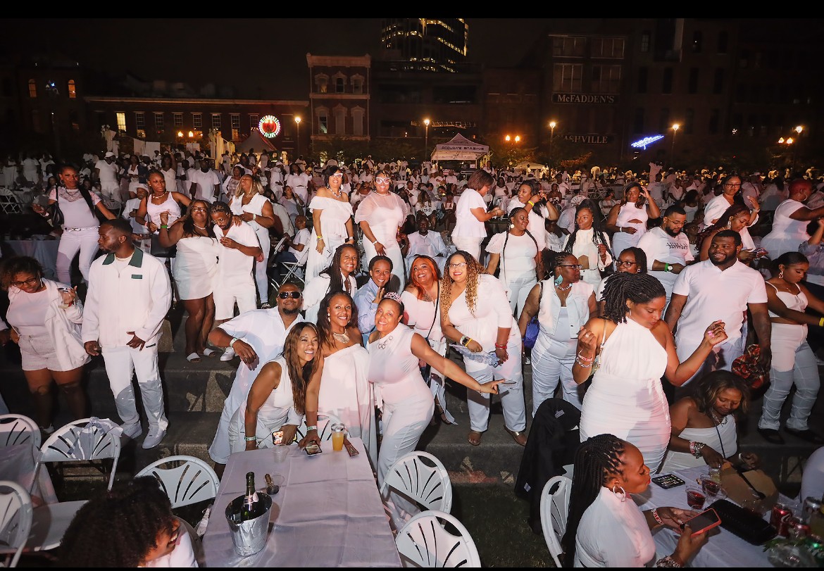 Crowd of people dressed in white gathered outdoors at night, some dancing and socializing, with tables and chairs in foreground.