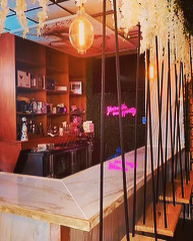Bar area with wooden shelves, hanging light, marble counter, and decorative elements, illuminated by warm lighting.