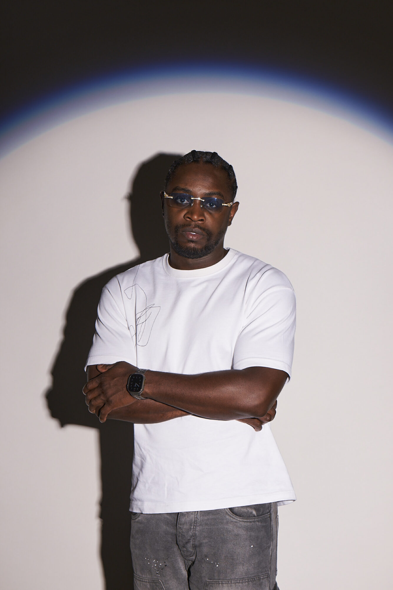 Man with sunglasses and a white t-shirt standing with arms crossed against a plain background, shadow visible.