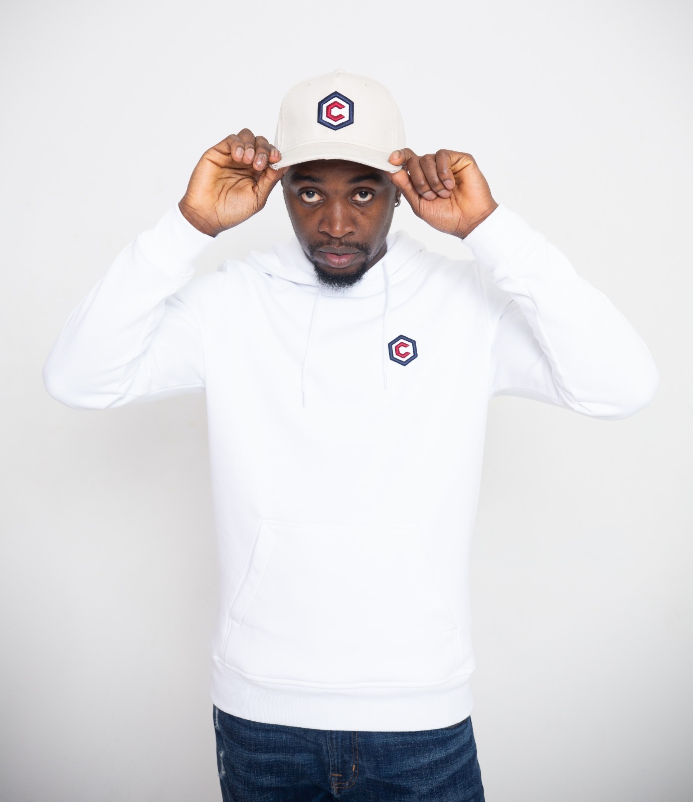 Man wearing a white hoodie and cap with a logo, adjusting the cap, standing against a plain white background.