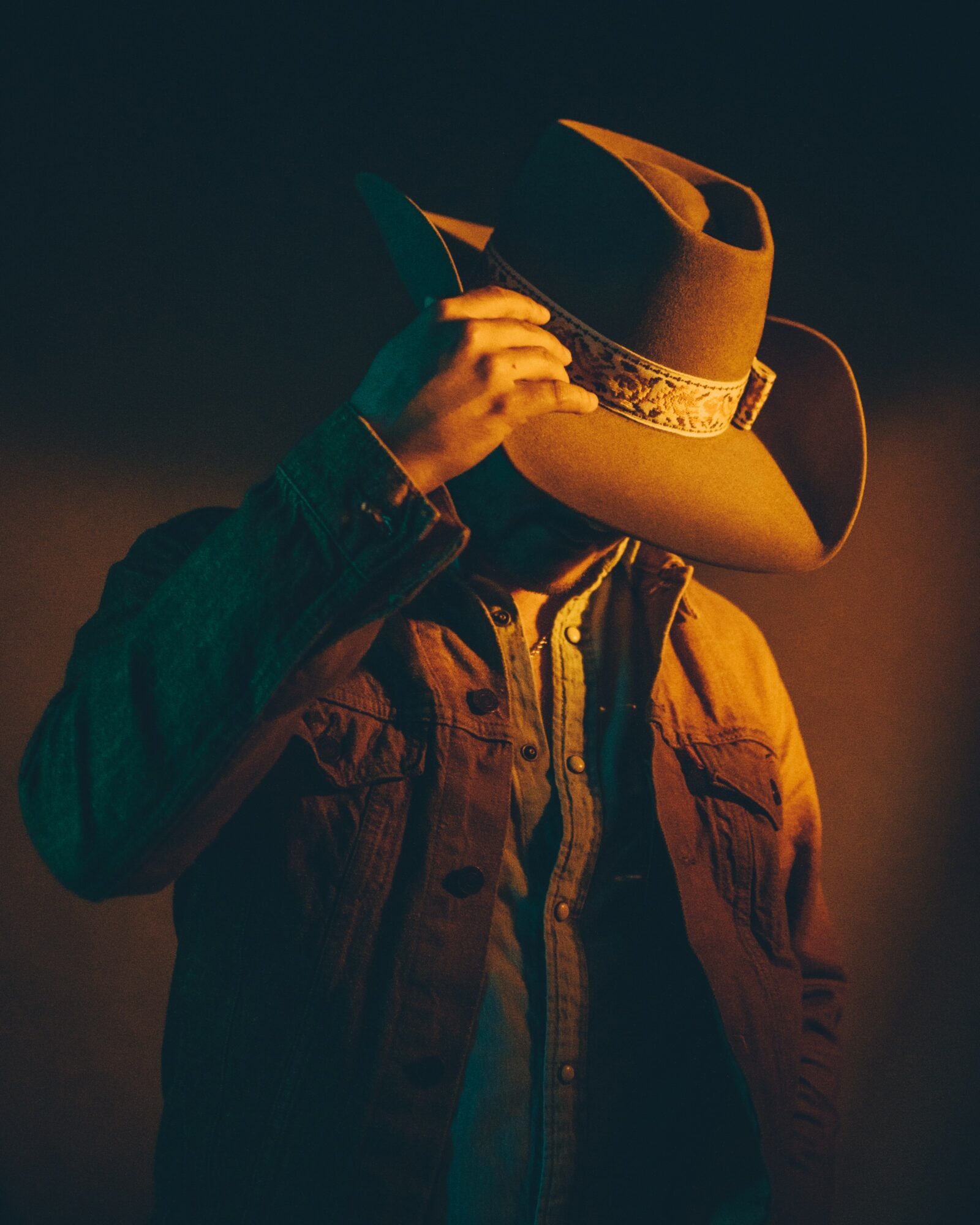 Person wearing a cowboy hat and denim jacket, adjusting the hat, with a dark background.