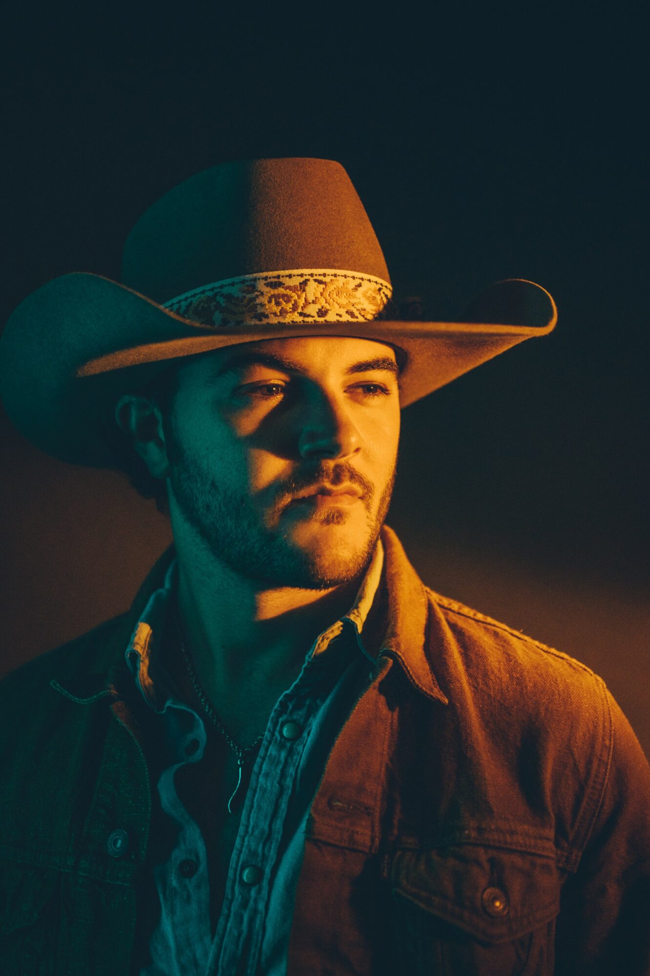 Man wearing a wide-brimmed hat and denim jacket, lit by warm light, against a dark background.