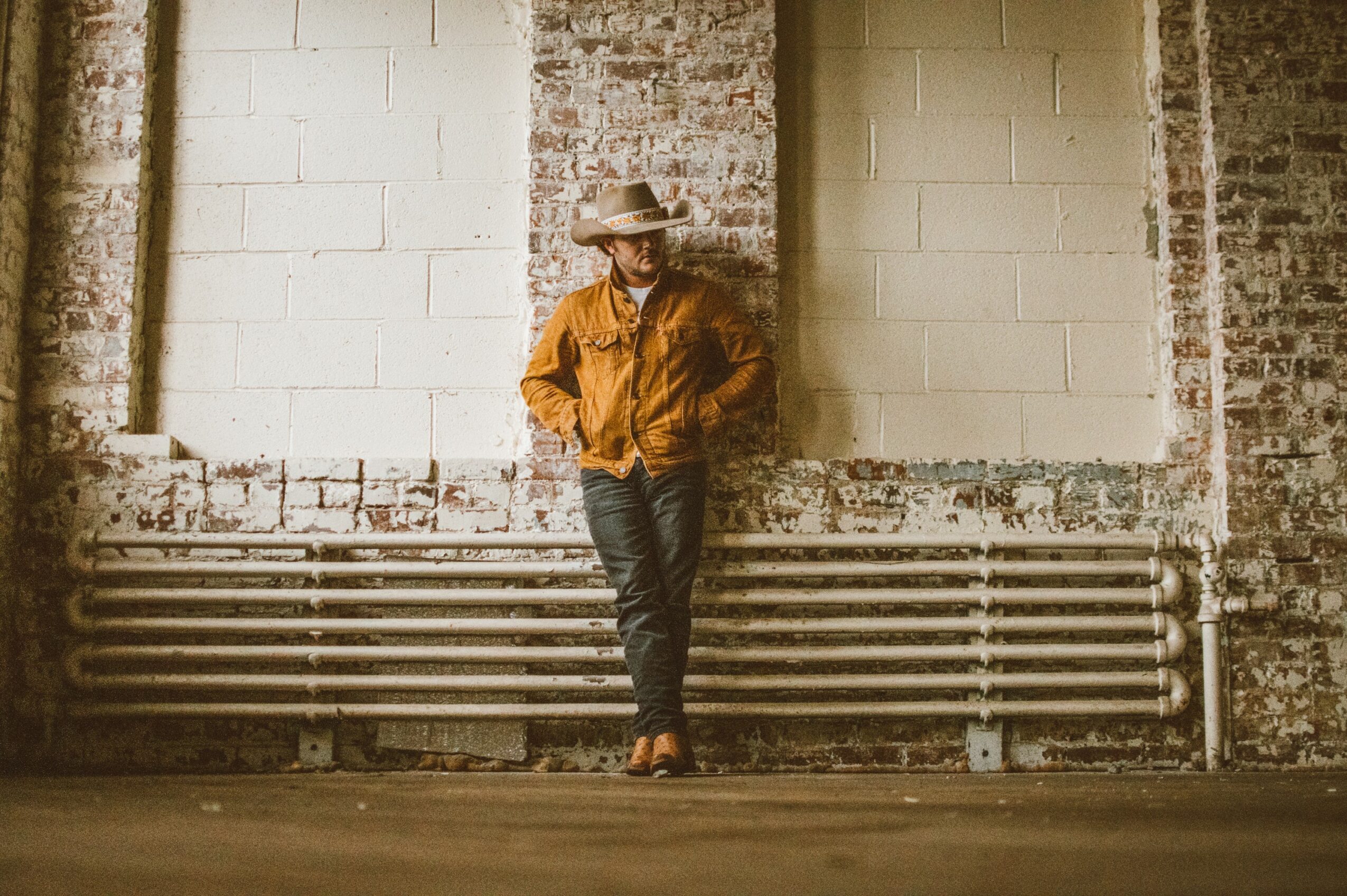 Person wearing a hat and orange jacket leaning against a wall in an industrial setting.