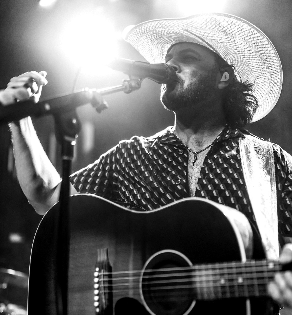 Man singing into microphone with guitar, wearing a wide-brimmed hat, in black and white, stage lighting in background.