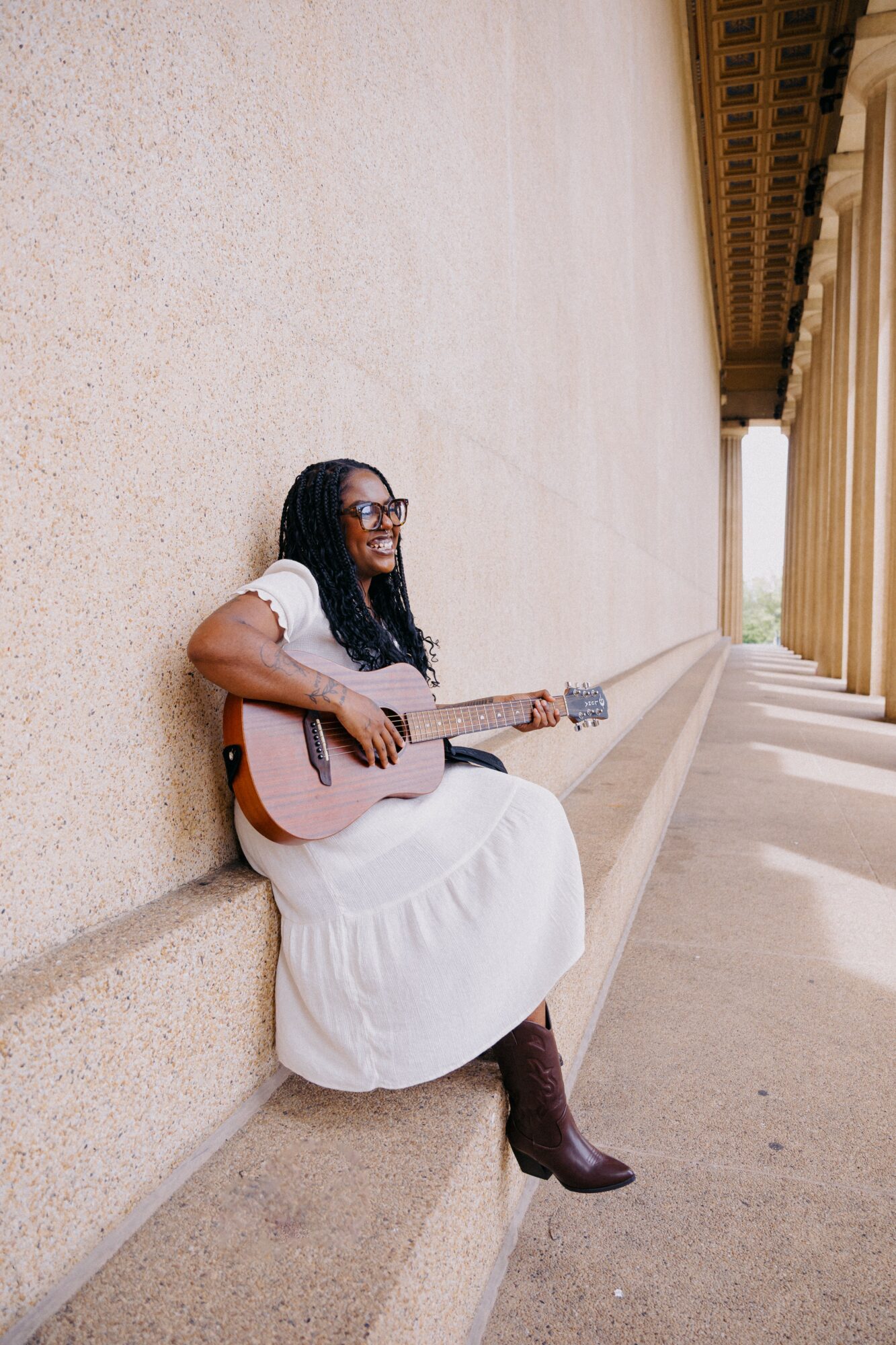 Woman sitting on a ledge playing guitar, wearing a white dress and boots, with columns in the background.