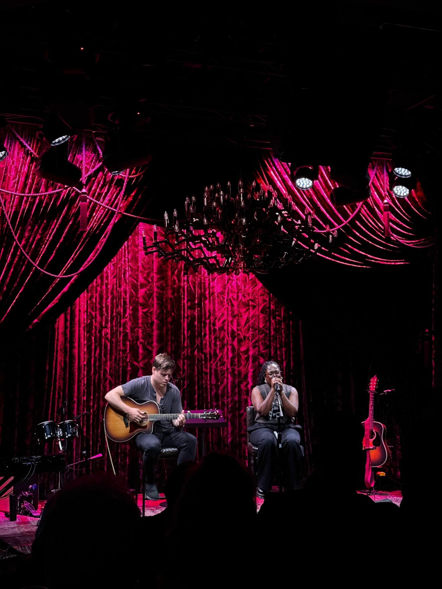 Musicians perform on stage with red curtains and pink lighting, playing guitars and singing, audience visible in foreground.