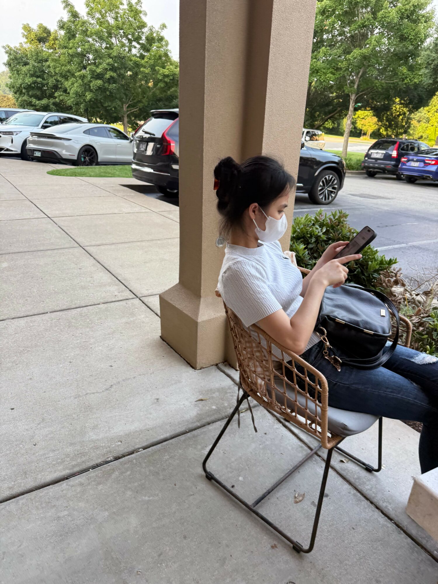 Person sitting on a chair outside, wearing a white shirt and face mask, holding a phone, with cars and trees in background.
