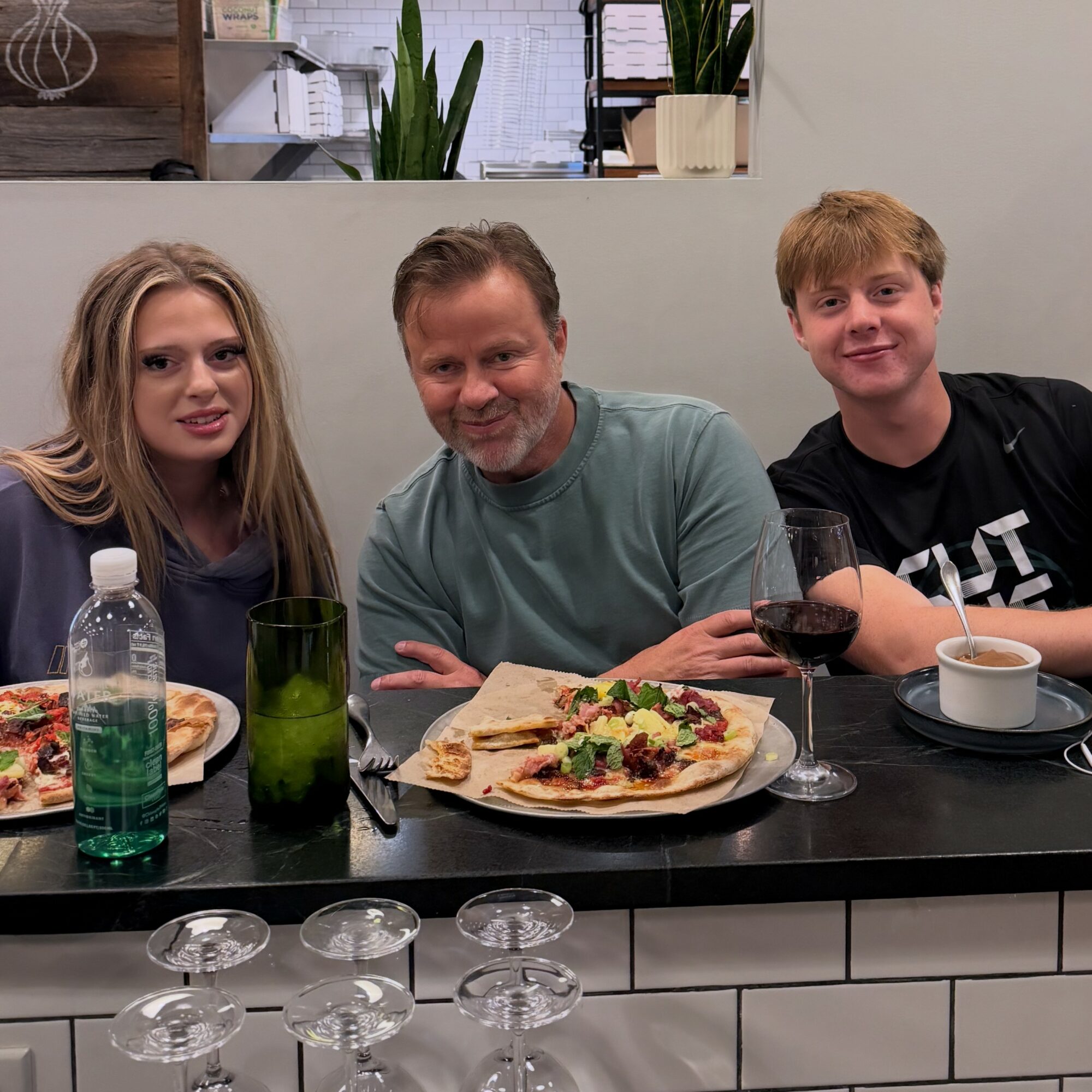 Three people sitting at a table with food and drinks, smiling, in a room with a window and plants behind them.