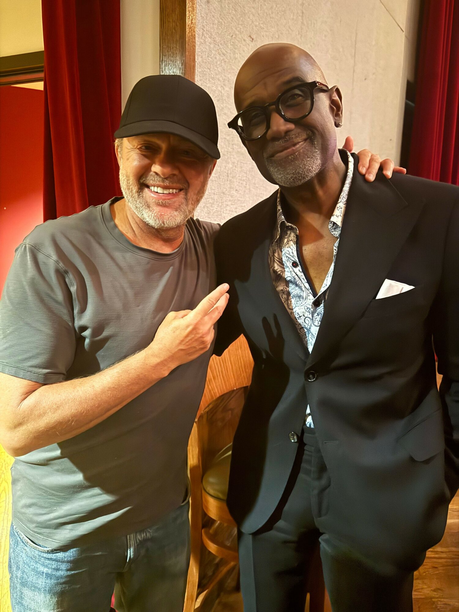 Two men standing indoors, one with glasses and a suit, the other with a cap and casual shirt, posing together.