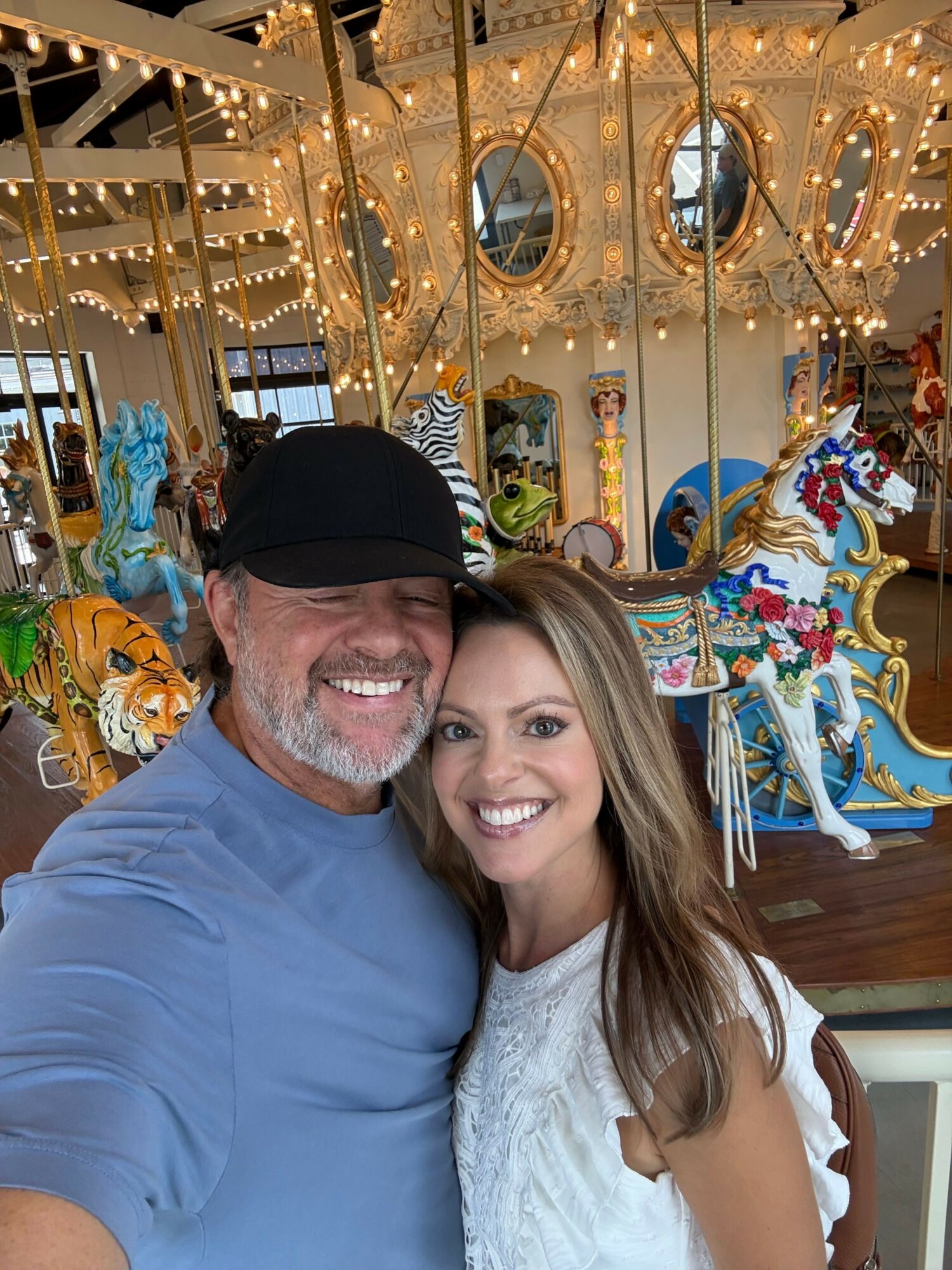 A smiling woman with long hair and a man with a beard and cap pose together indoors, with festive decorations in the background.