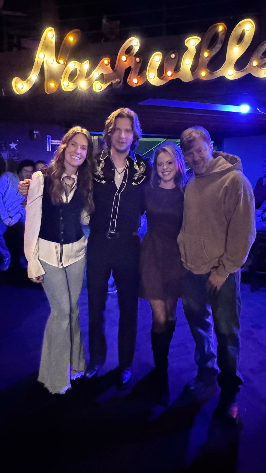 Four people standing together in front of a neon sign that reads 'Nashville' at night.
