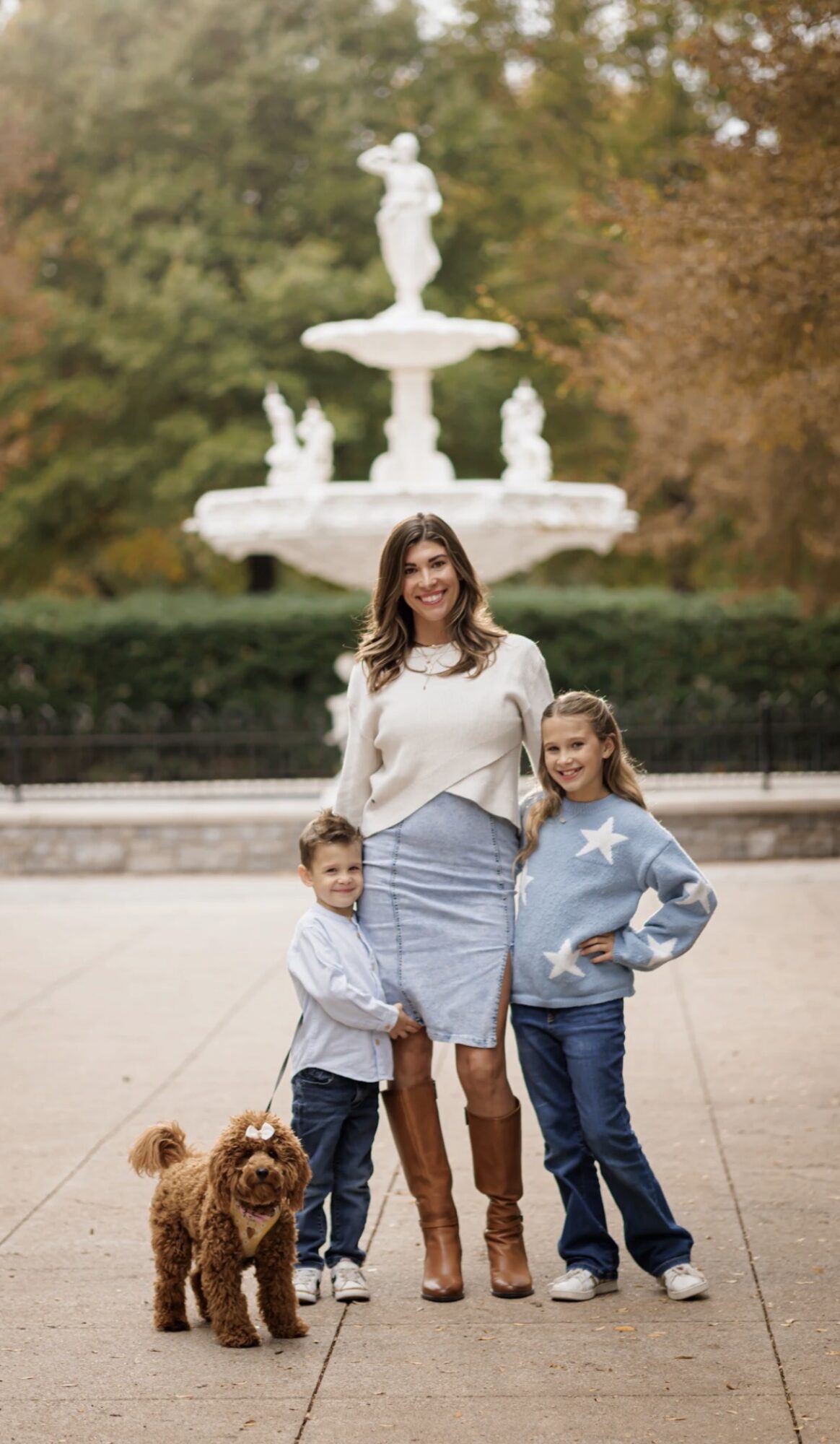 Woman with two children and a dog in front of a fountain with statues, outdoors, trees in background.