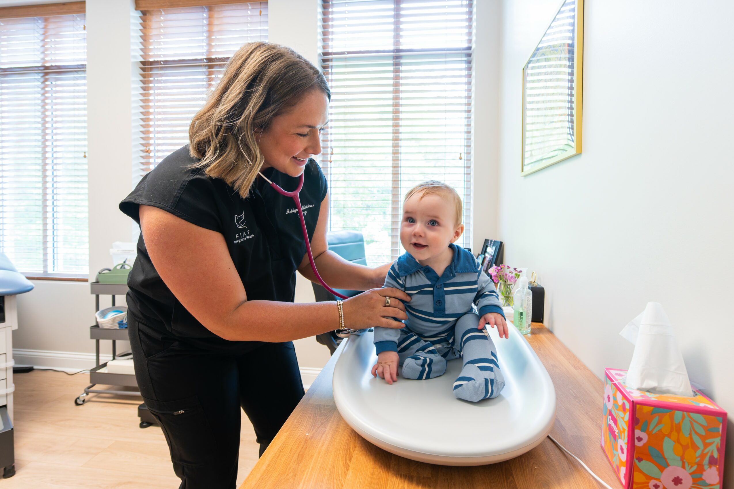 A woman using a stethoscope to listen to a baby's chest on a changing table in a bright room.