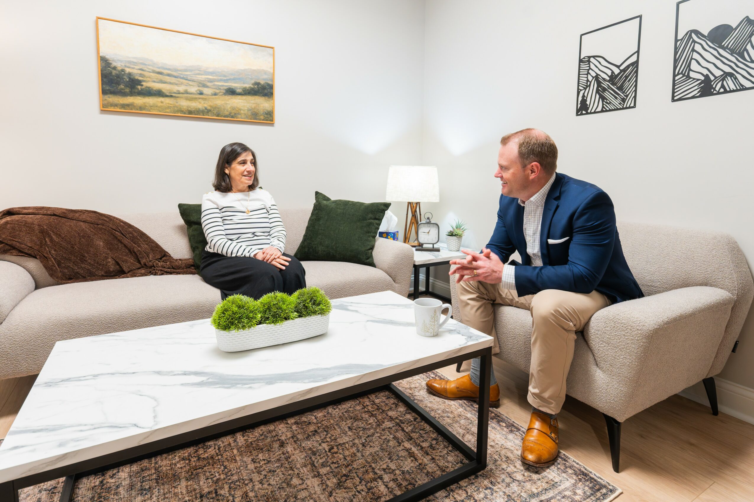 Two people sit on sofas in a living room, engaged in conversation, with artwork and a lamp on the wall behind them.