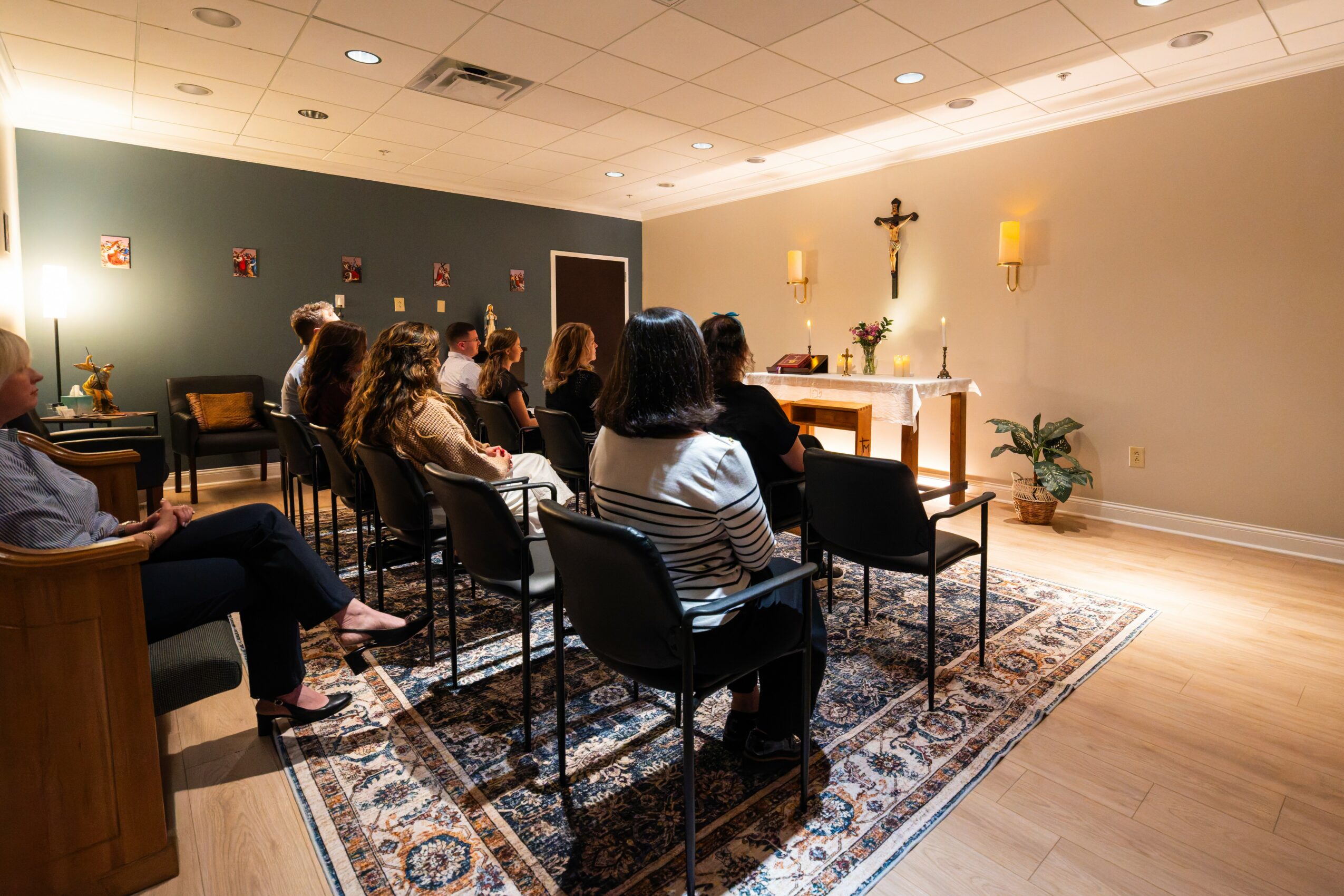 People seated in chairs facing an altar with a cross and candles in a room, possibly a prayer or meditation session.