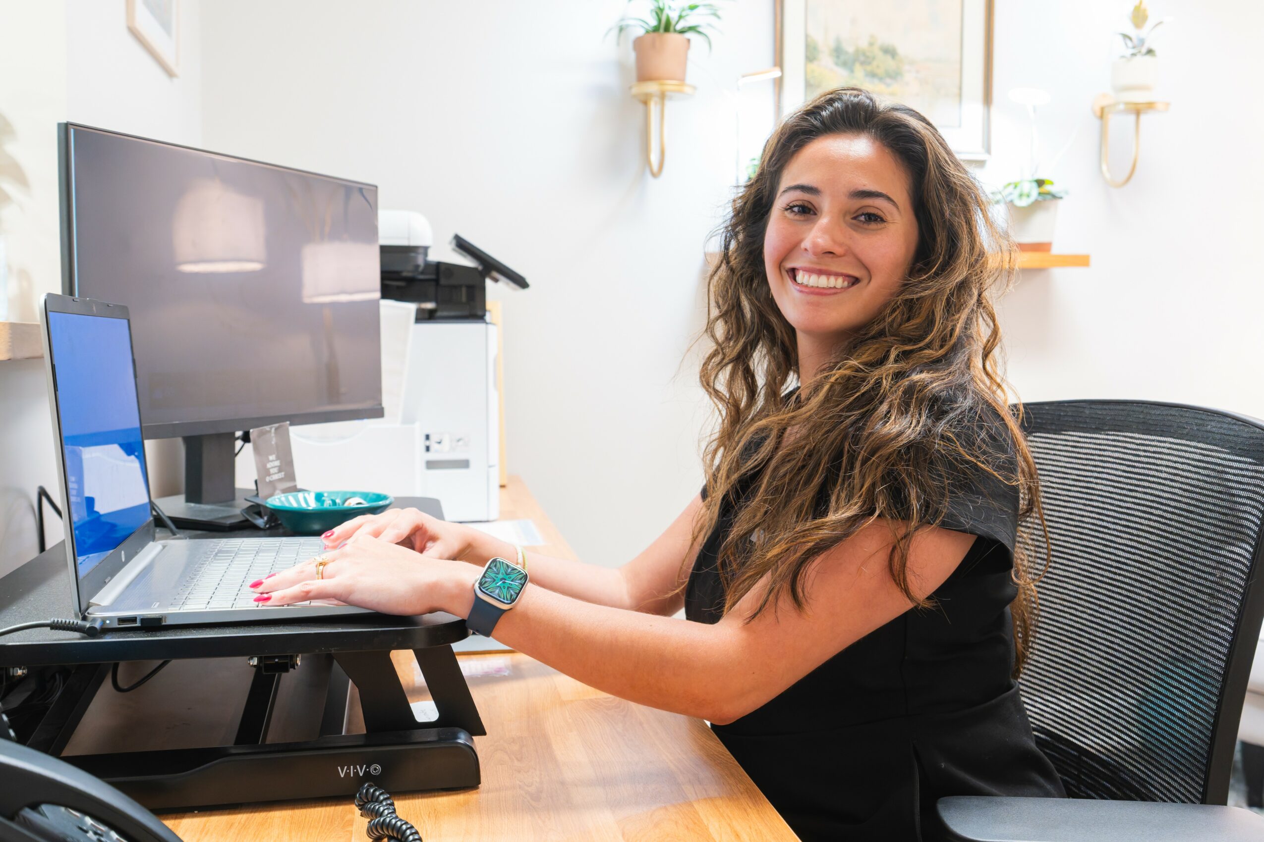 Smiling woman with long curly hair working at a desk with multiple monitors in a bright room.