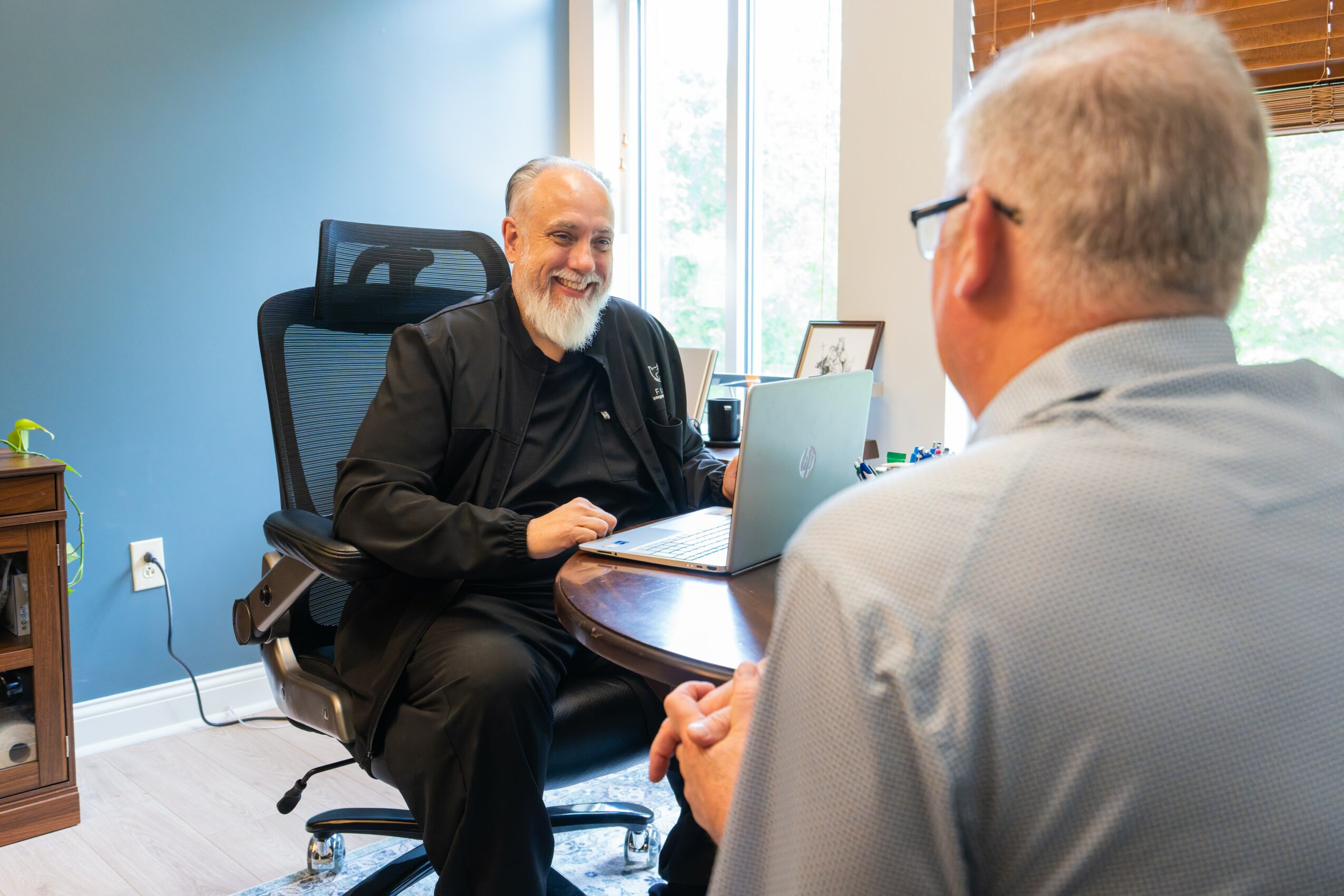 Two men are having a conversation in an office, one is smiling and sitting at a desk, the other is facing away.