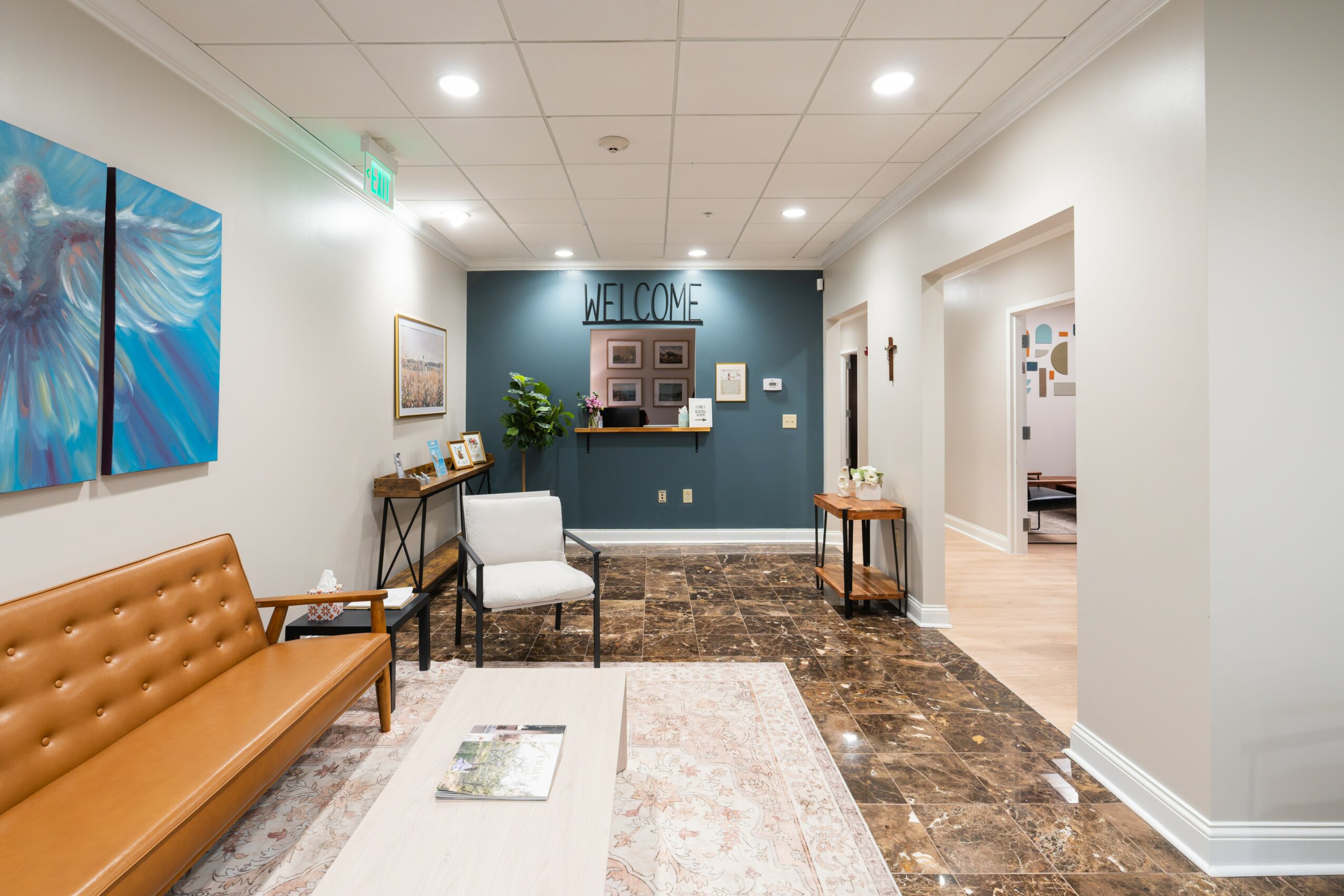 Lobby with brown sofa, white chair, artwork, and a welcome sign on a blue wall, with a doorway to another room.