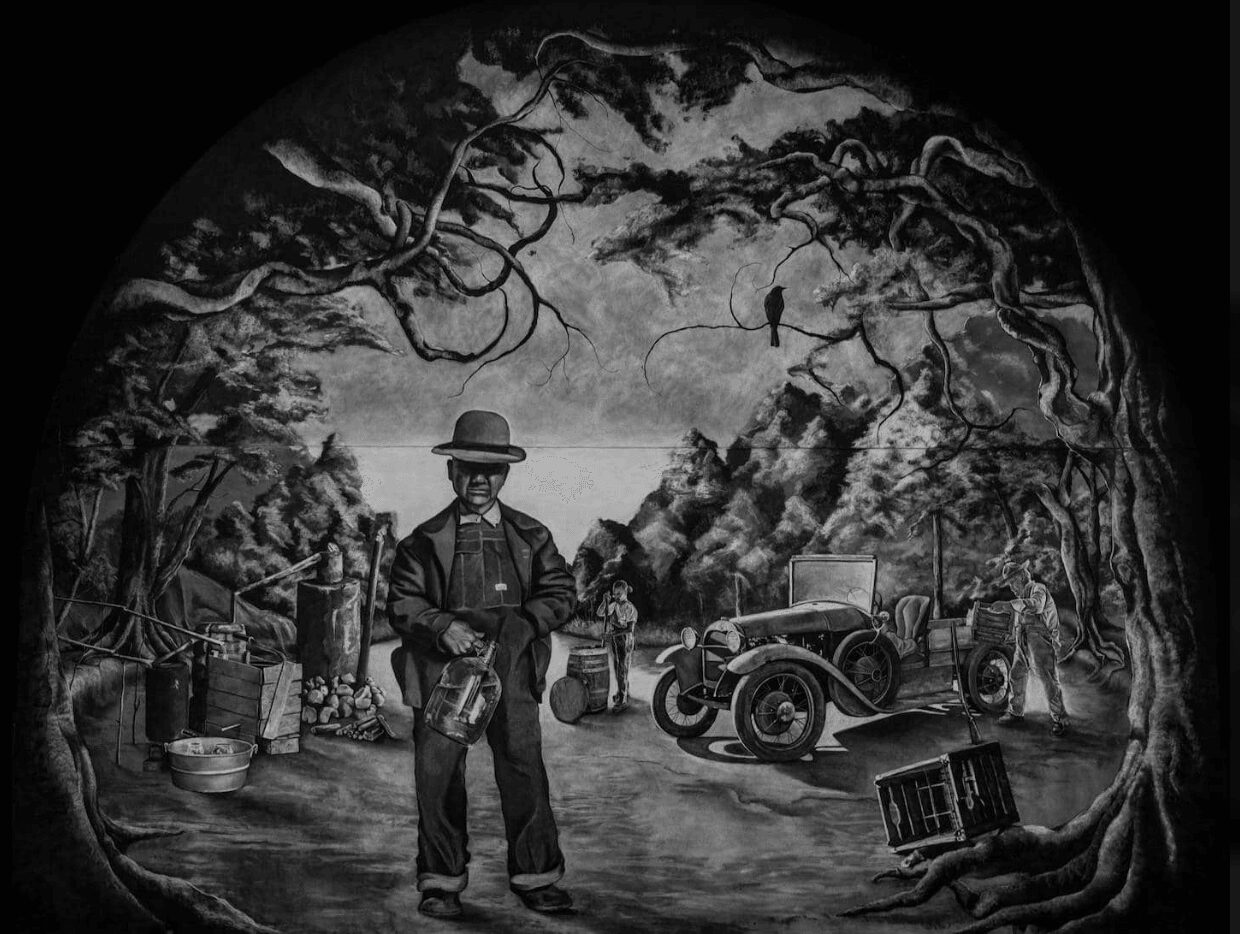 Black and white scene of a man in a hat and coat standing outdoors near a vintage car, with trees and a river in the background.