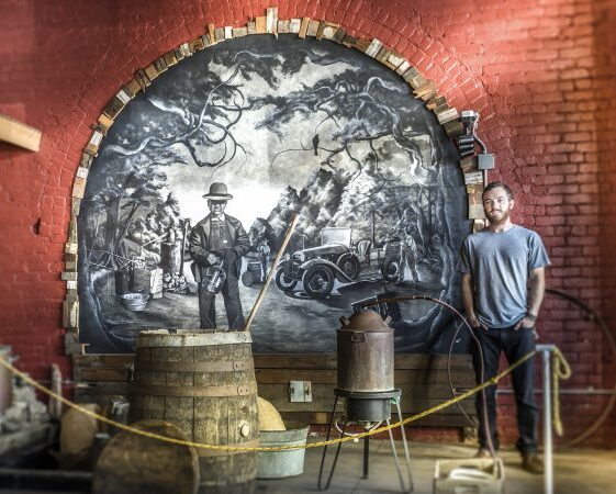 Two people stand in front of a large circular mural depicting a historical scene with a man, trees, and a vintage car, inside a brick-walled space.