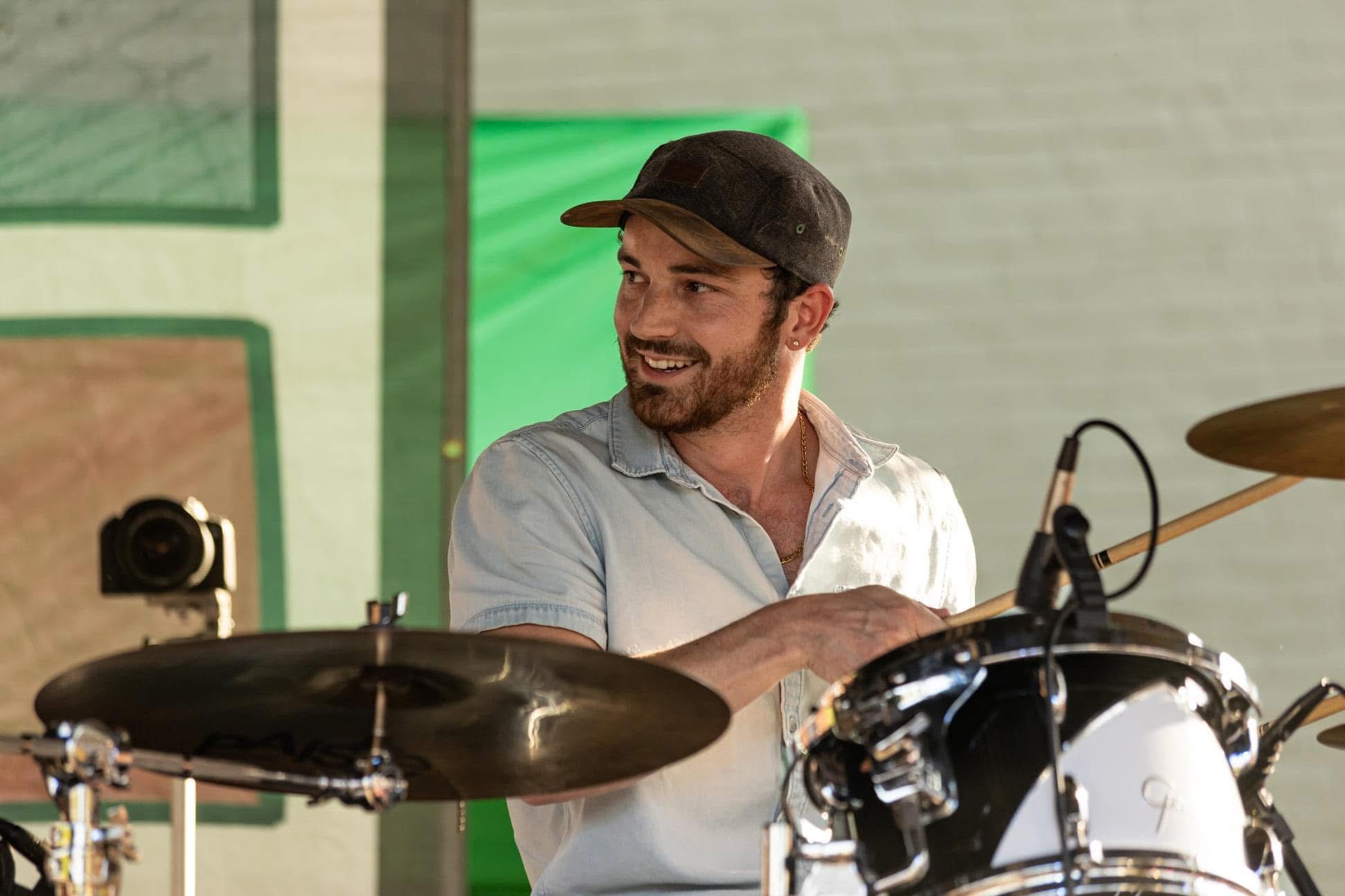 Man with a beard wearing a cap and white shirt playing drums, smiling, in a room with green and beige walls.