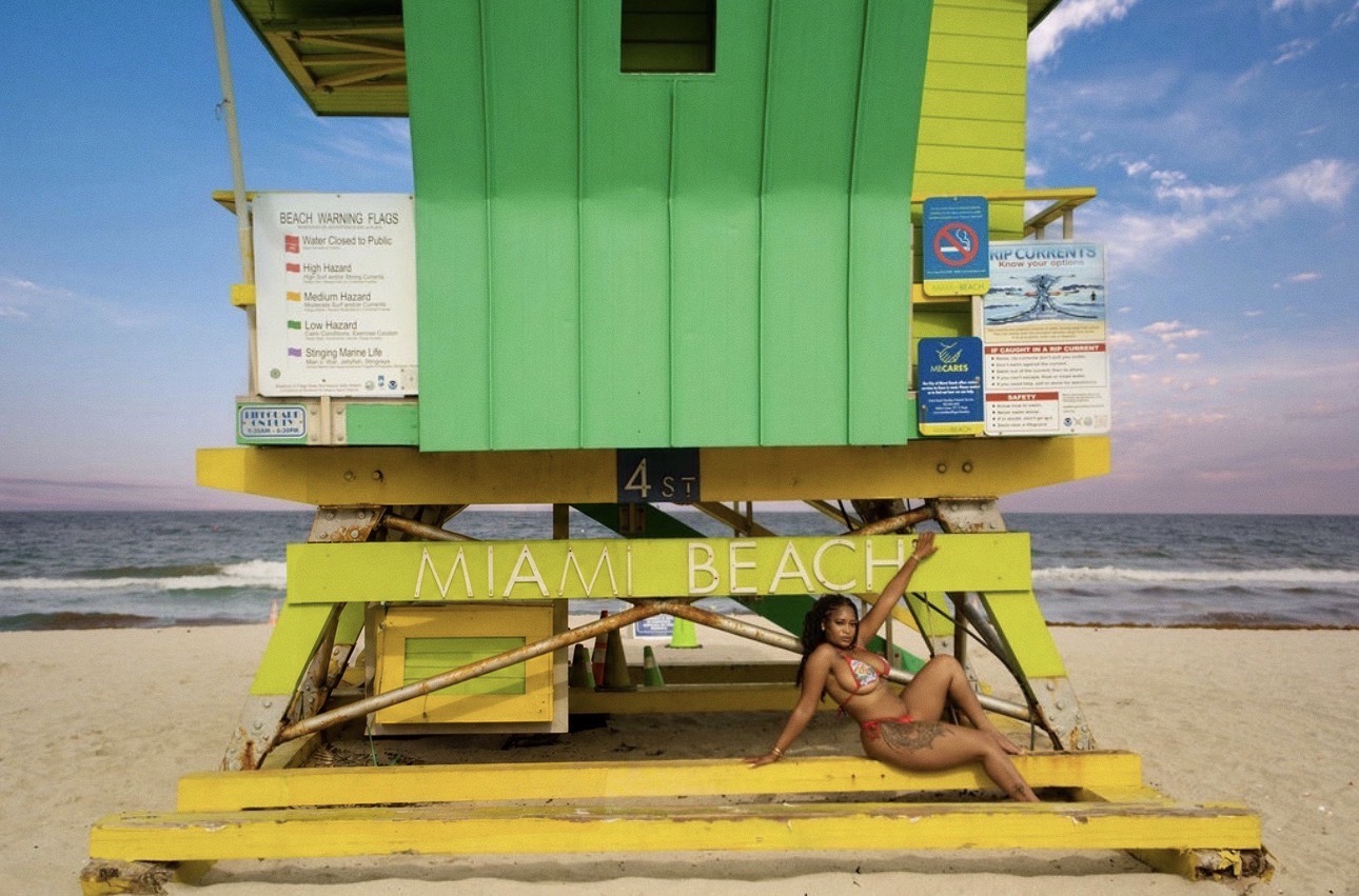 Person lying on a yellow bench in front of a colorful lifeguard tower on the beach, ocean and sky in background.