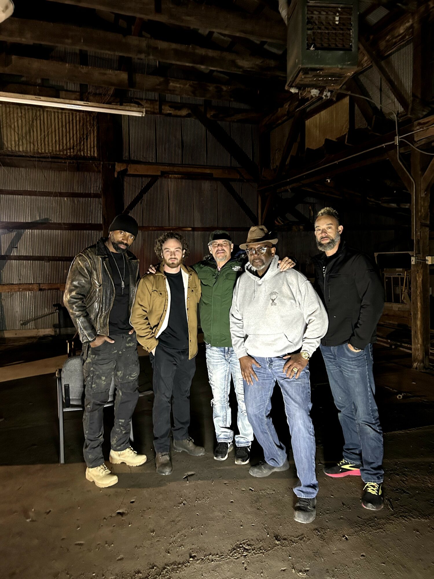 Five people standing inside a rustic wooden building, some wearing jackets and casual clothing, posing for a photo.