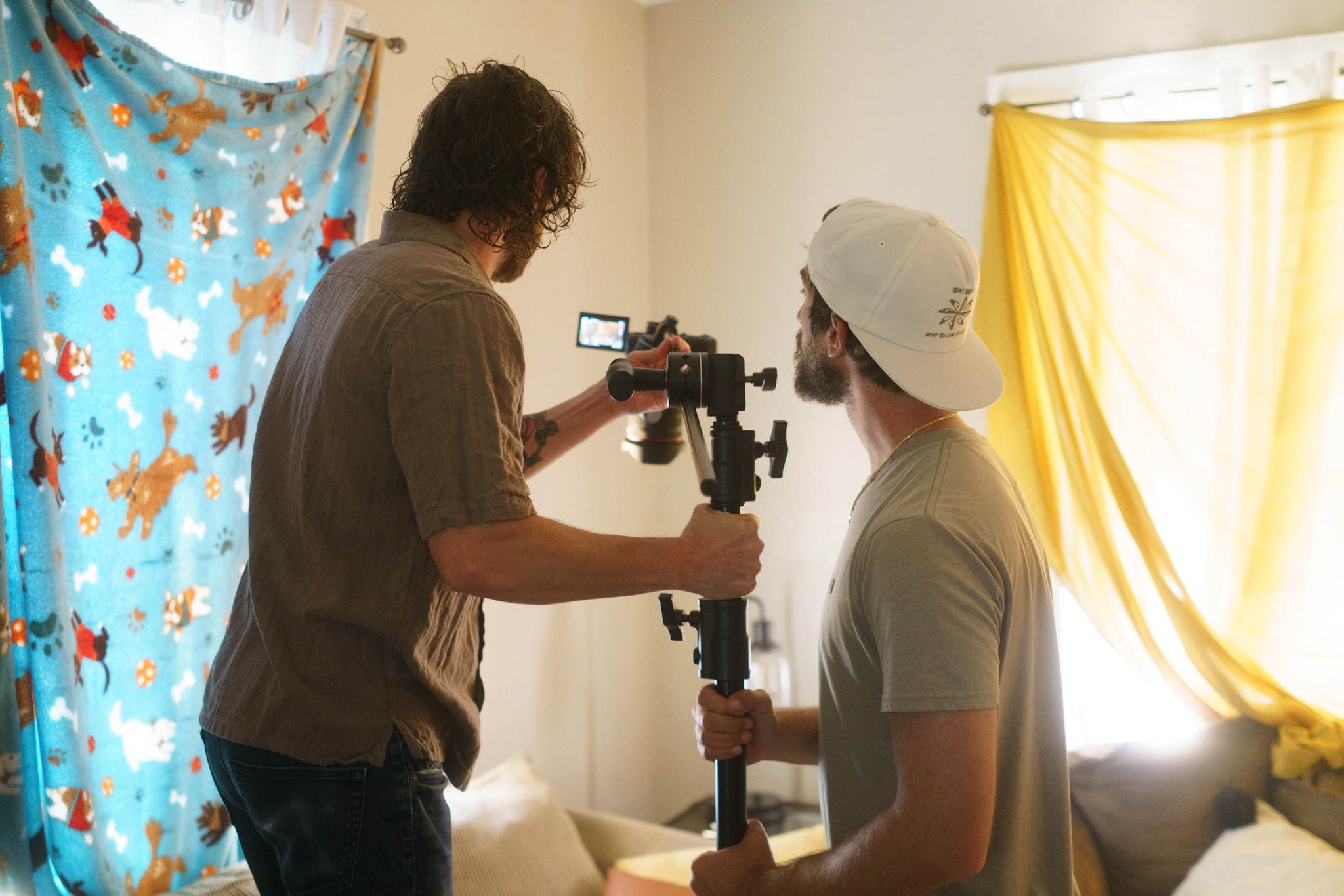 Two men filming indoors, one holding a camera stabilizer, sunlight coming through yellow curtains, colorful backdrop on wall.