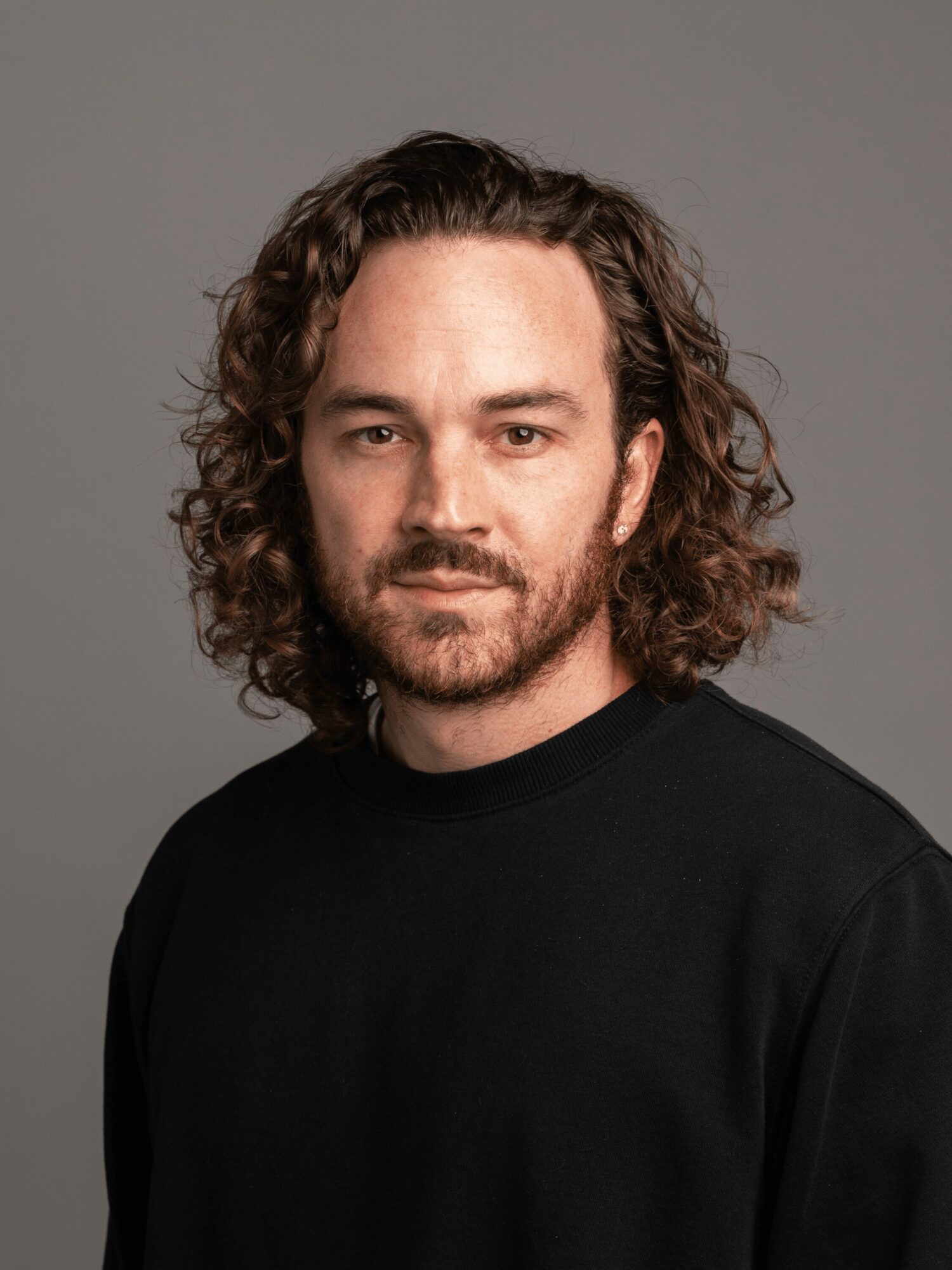 Man with curly hair and beard wearing a black shirt, looking at the camera against a gray background.